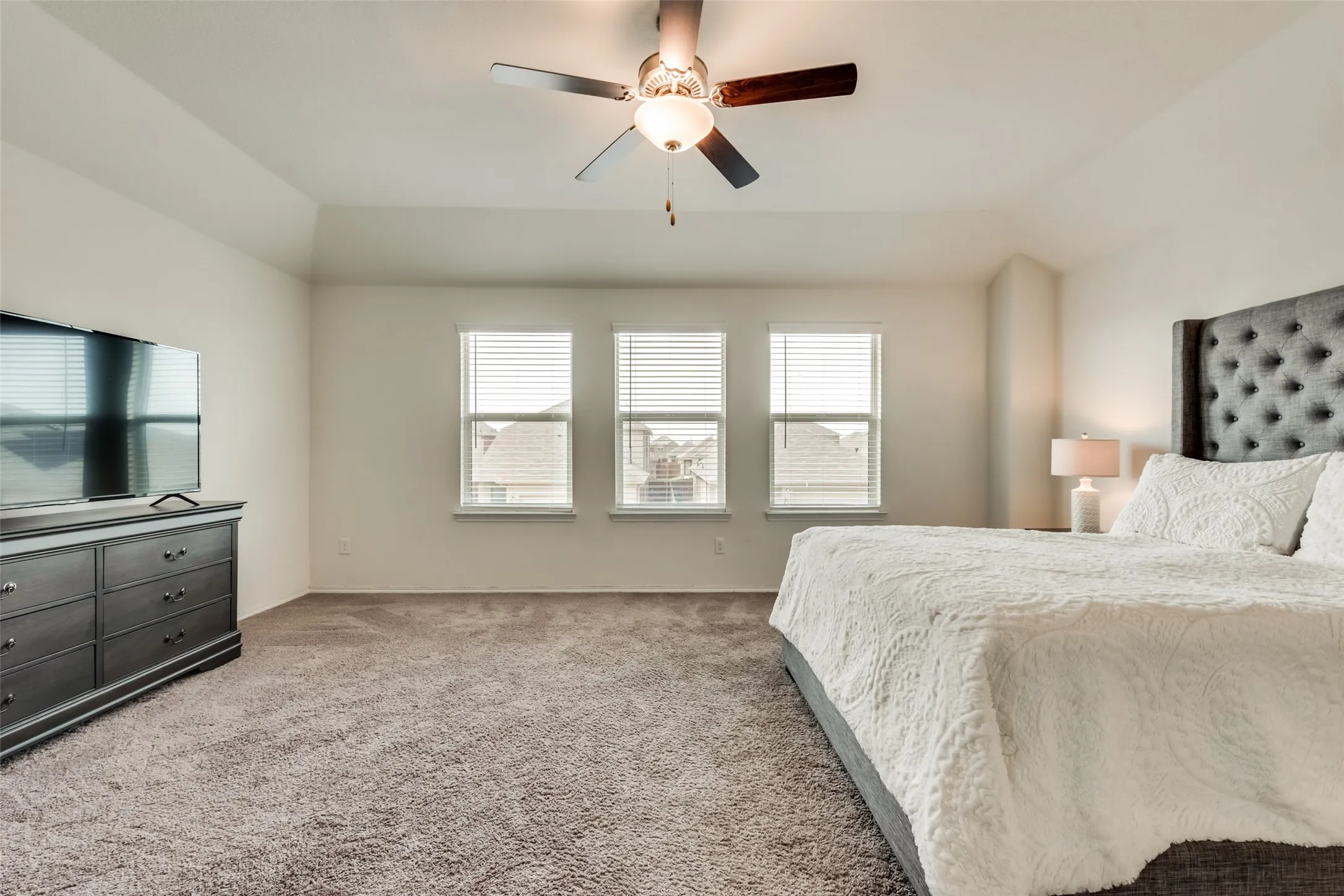 Spacious room featuring vaulted ceilings, neutral-toned carpet, three windows with blinds, and a ceiling fan with integrated lighting