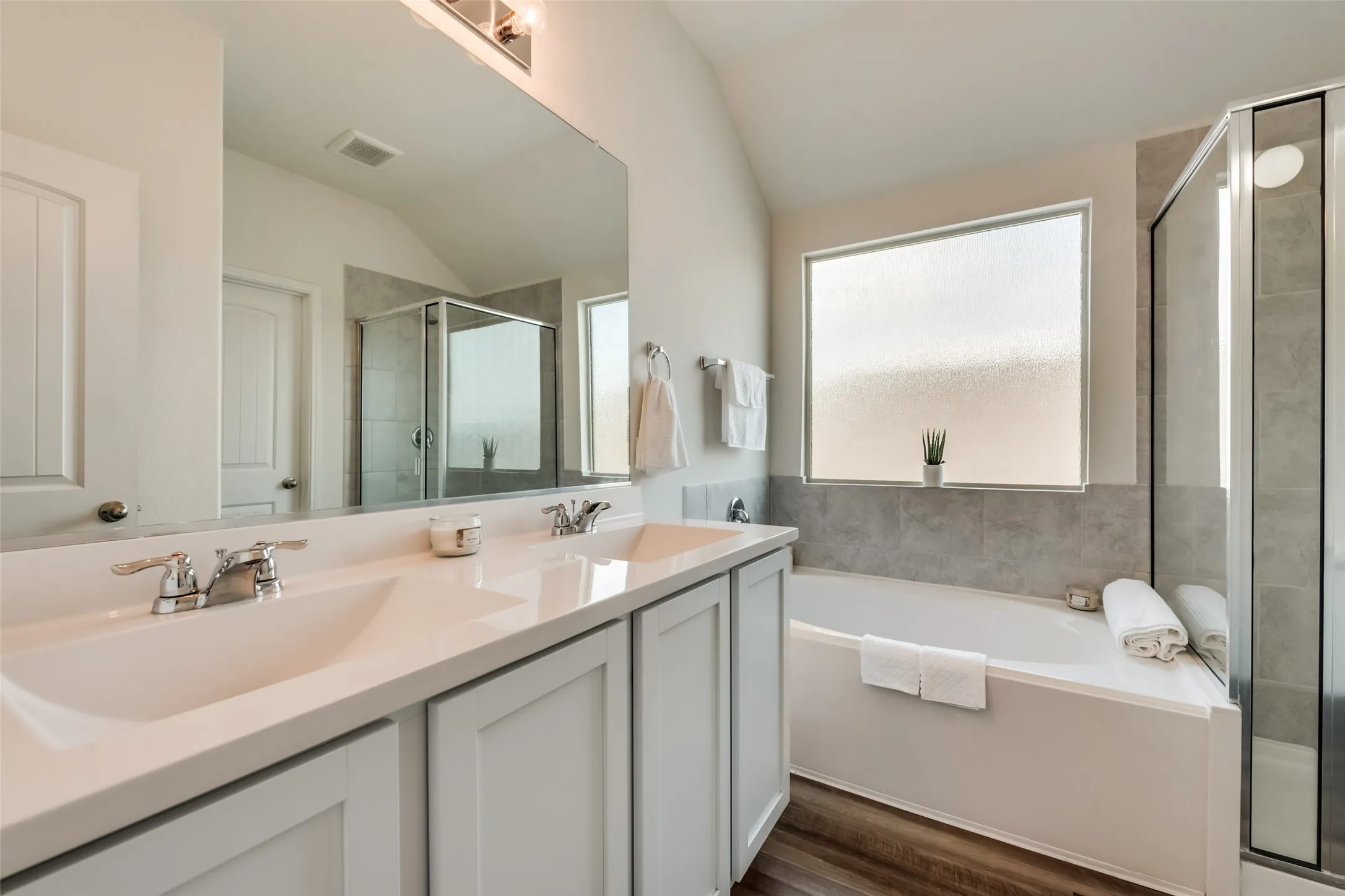 Bathroom featuring a double vanity with light-toned cabinetry, a built-in soaking tub, a separate glass-enclosed shower, wood-finish flooring, and a large privacy window