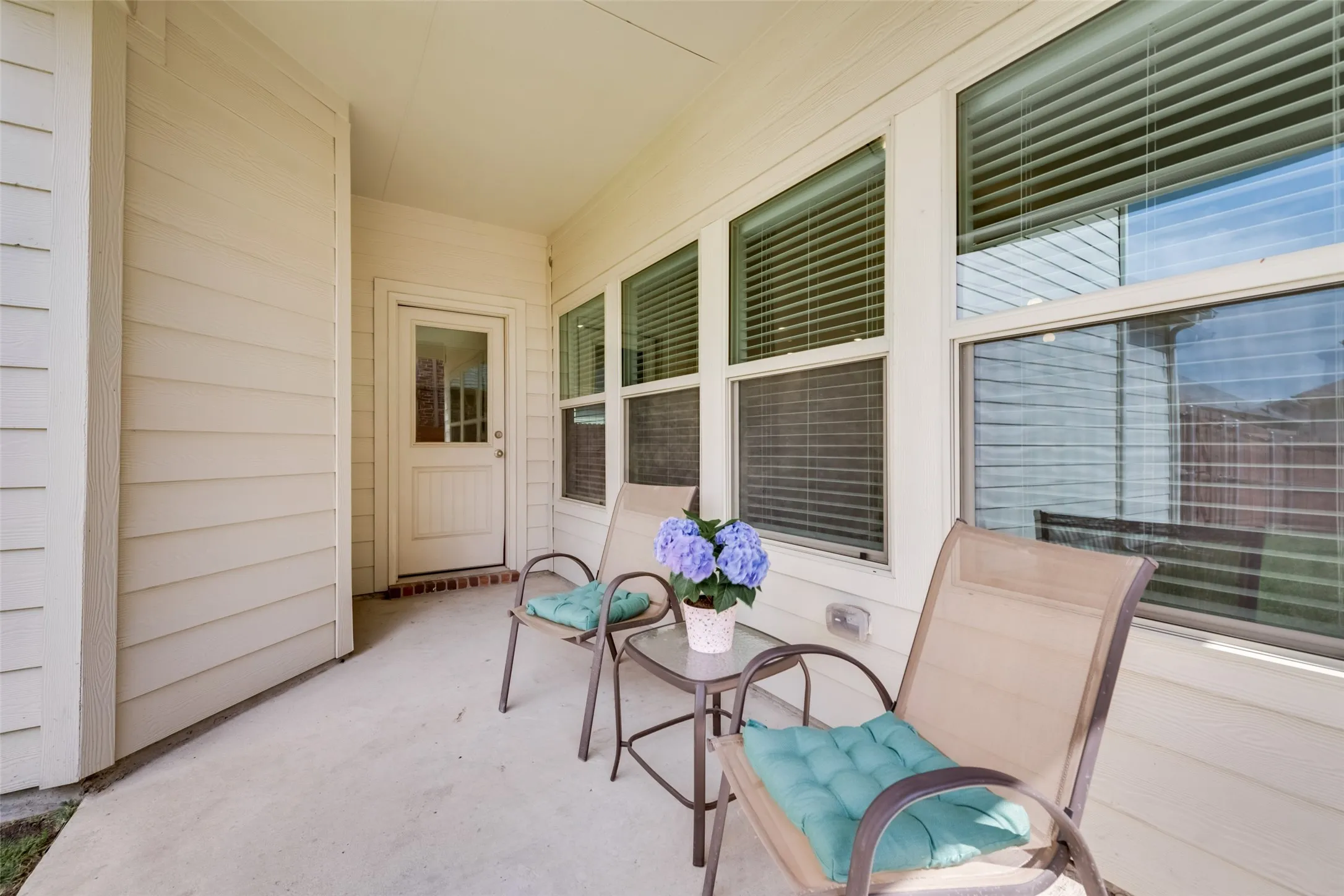 Covered concrete patio with horizontal siding, multiple windows featuring blinds, and a glass-paneled exterior door