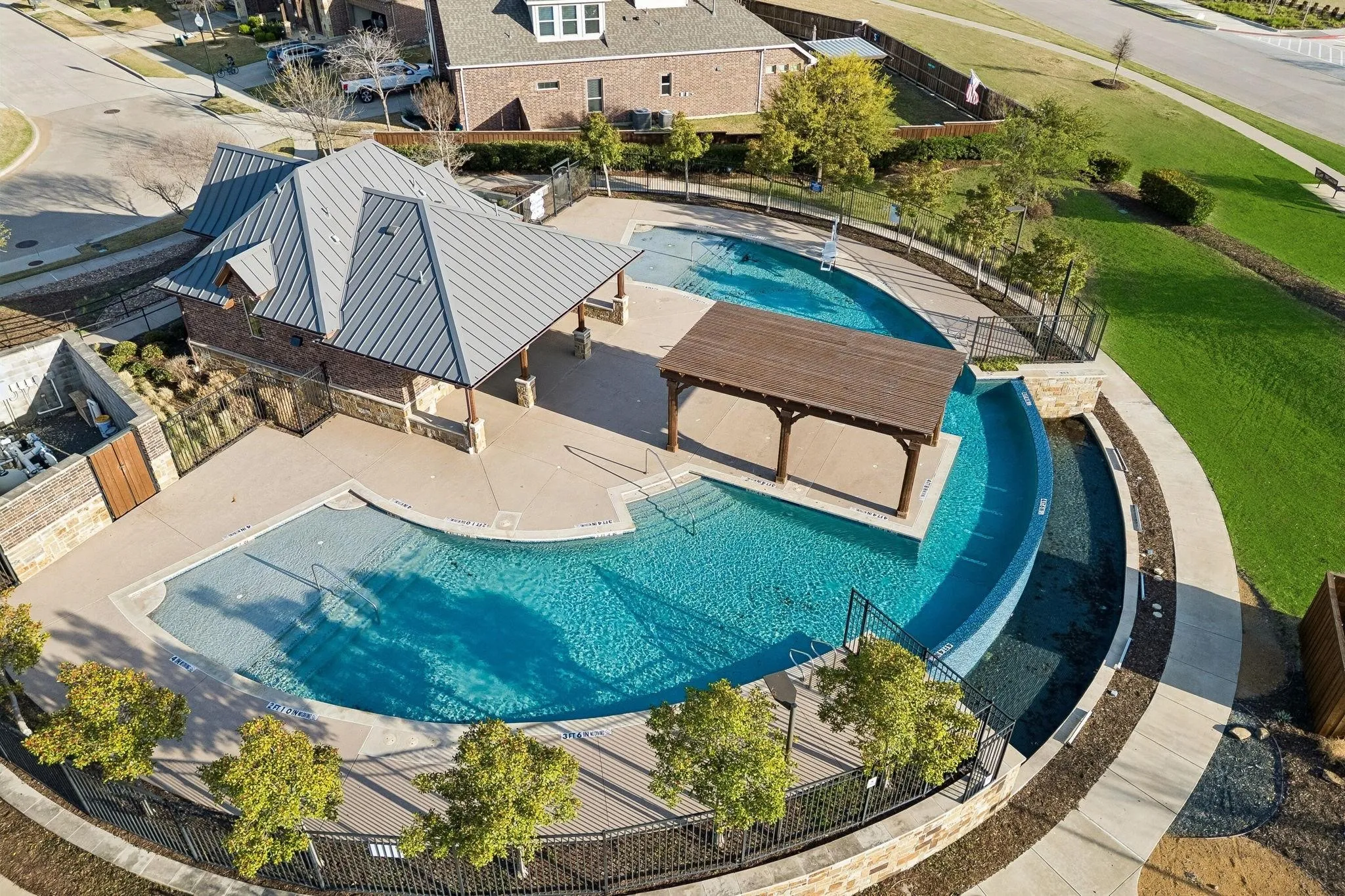 Curved swimming pool featuring a pavilion with a metal roof, an in-pool pergola, surrounding concrete decking, and adjacent green spaces