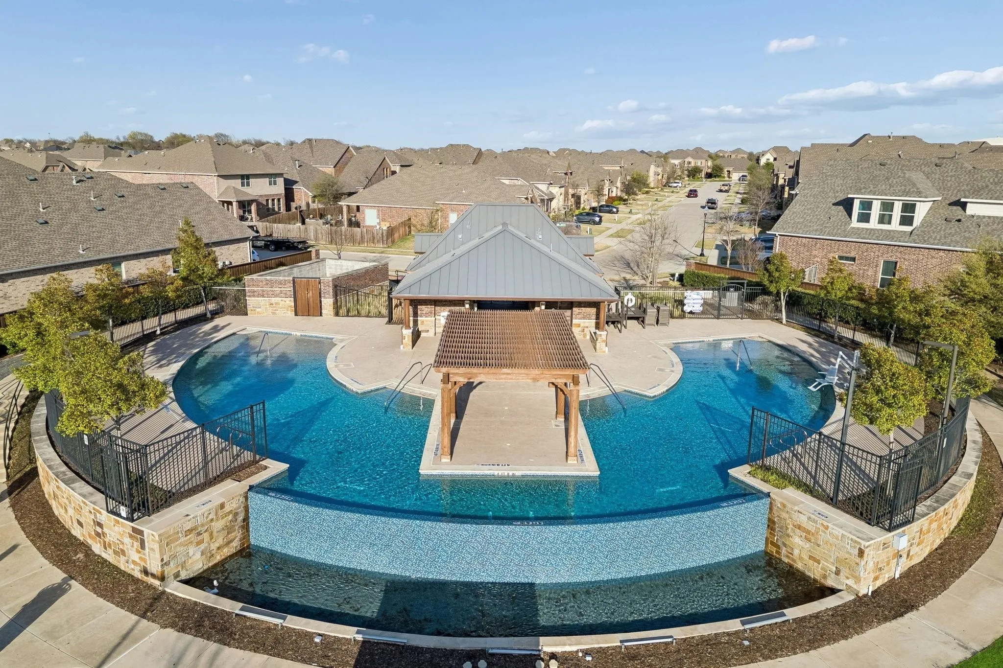 Community pool area featuring a multi-tiered swimming pool, a central covered seating area, a stone-clad retaining wall with a tiled water feature, a main building with a metal roof, and surrounding paved walkways