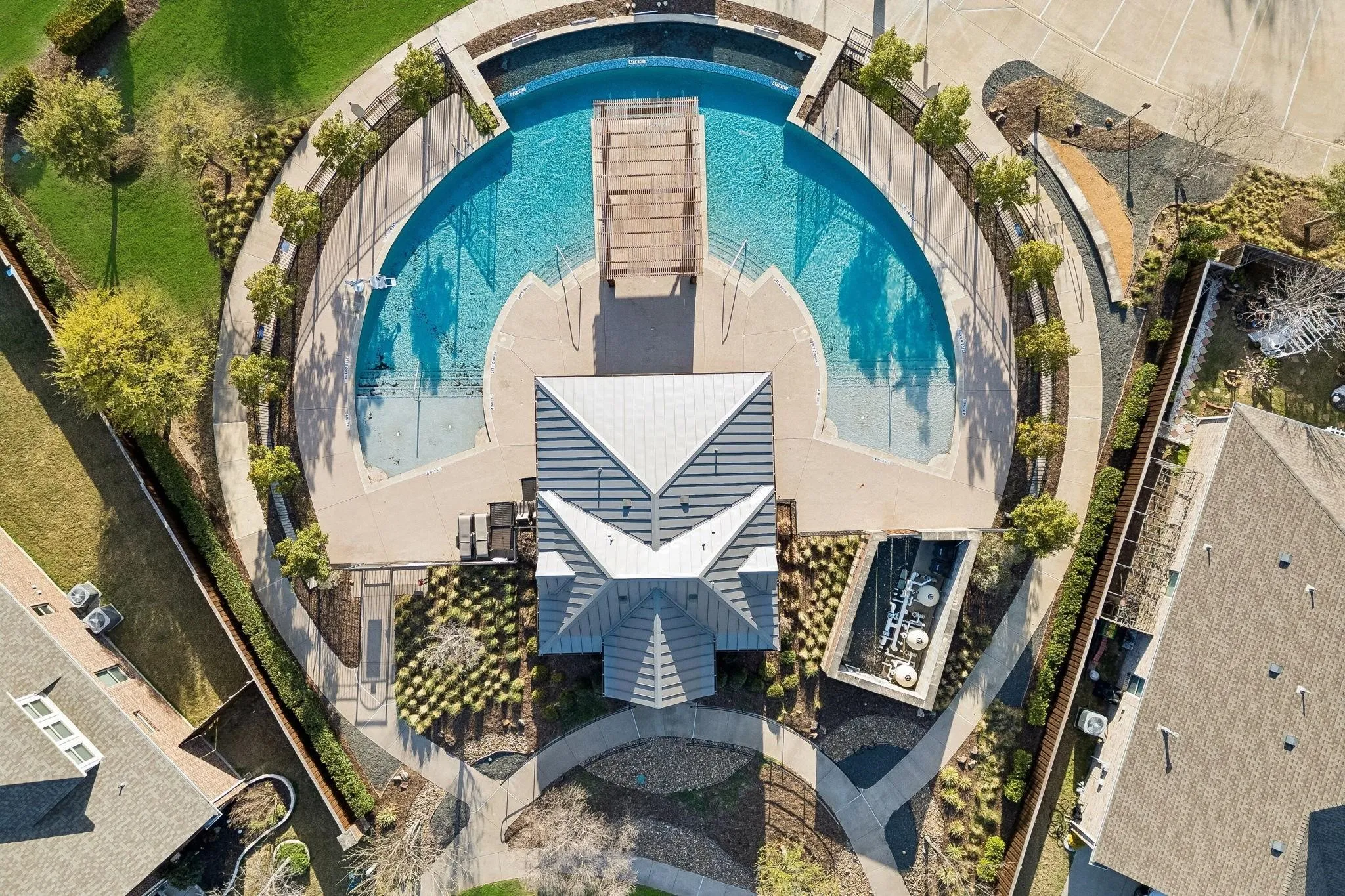 Community pool with integrated wading areas, surrounded by concrete decking, a central pavilion with a metal roof, and manicured landscaping