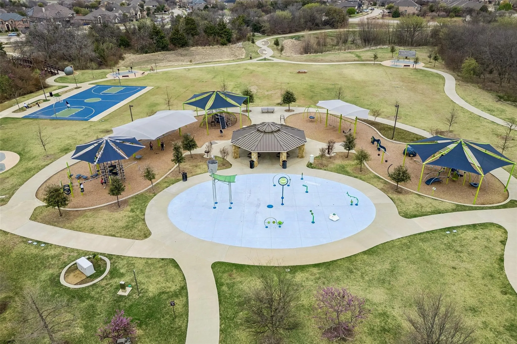 Expansive park grounds featuring a splash pad, multiple shaded play areas, a covered pavilion, a sports court, and meandering paved pathways