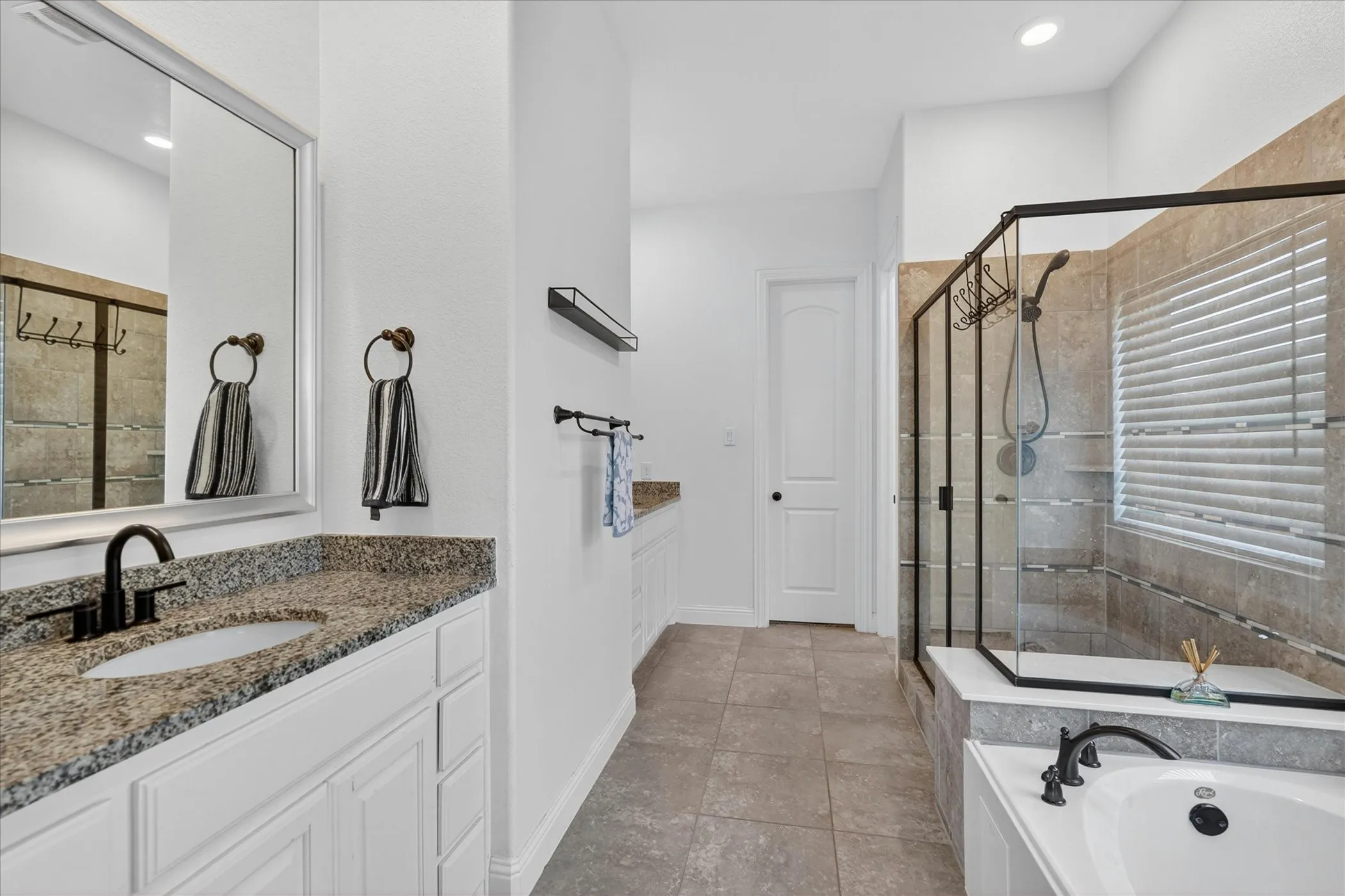 Bathroom featuring a spacious walk-in shower with clear glass enclosure and tiled walls, a separate built-in bathtub, dual vanity sinks with granite countertops and white cabinetry, and large format tile flooring