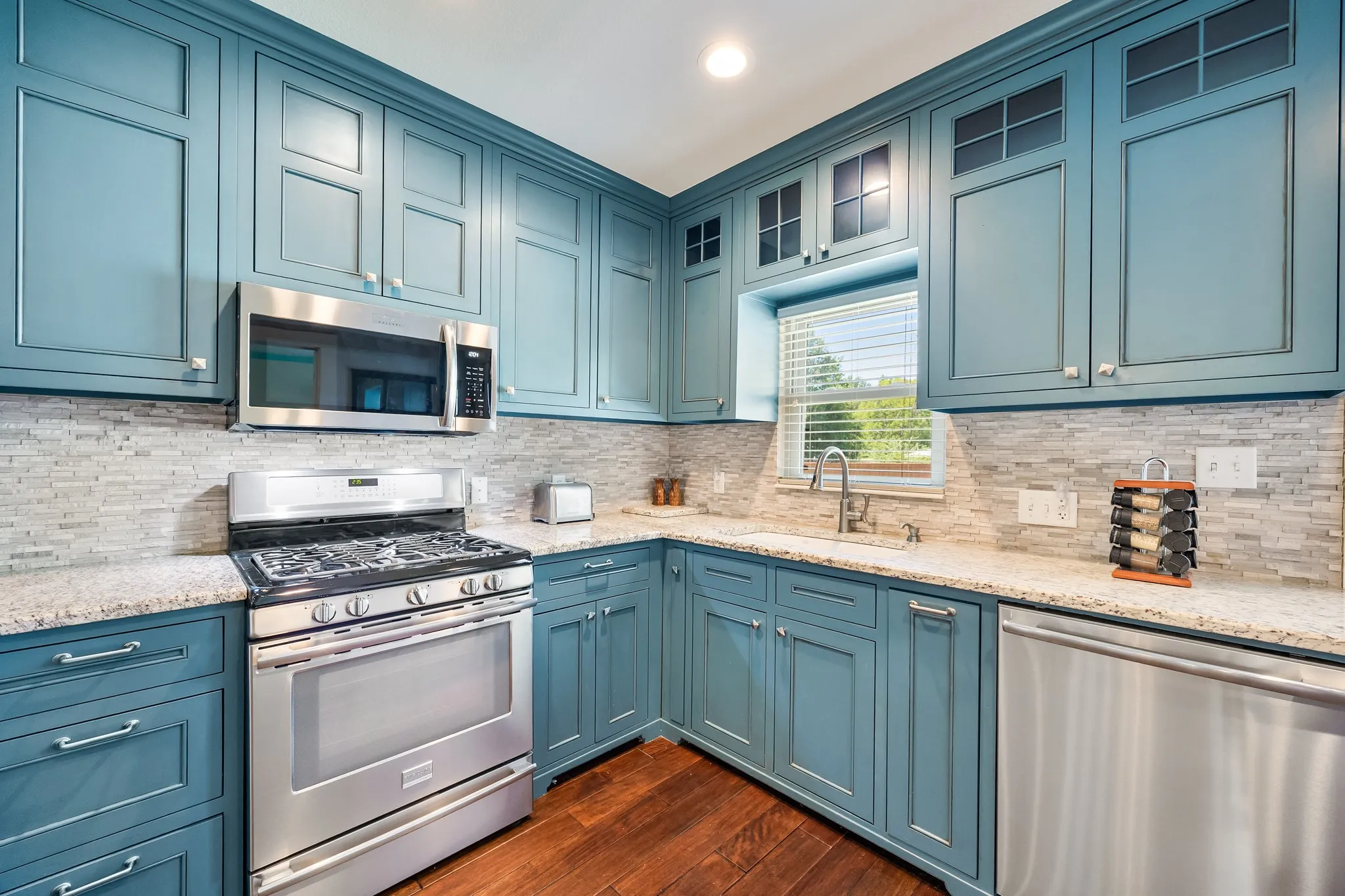 Kitchen featuring blue shaker-style cabinetry with upper glass-front accents, stainless steel appliances, speckled countertops, and a full-height tile backsplash