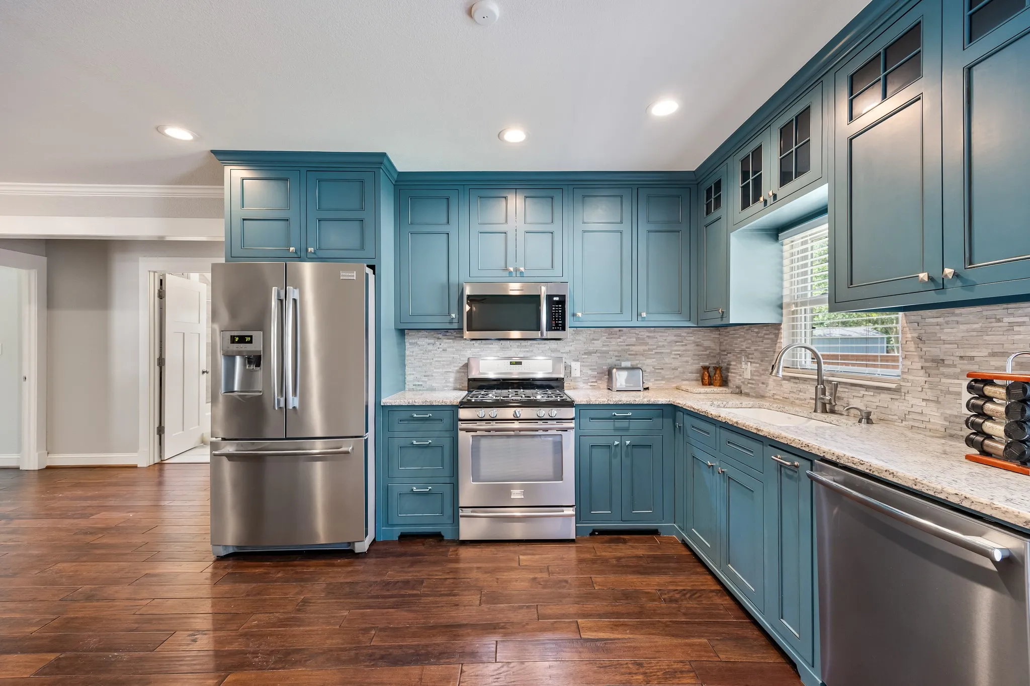 Kitchen featuring wood-finish flooring, teal cabinetry, stainless steel appliances, light-toned stone countertops, and a full tile backsplash