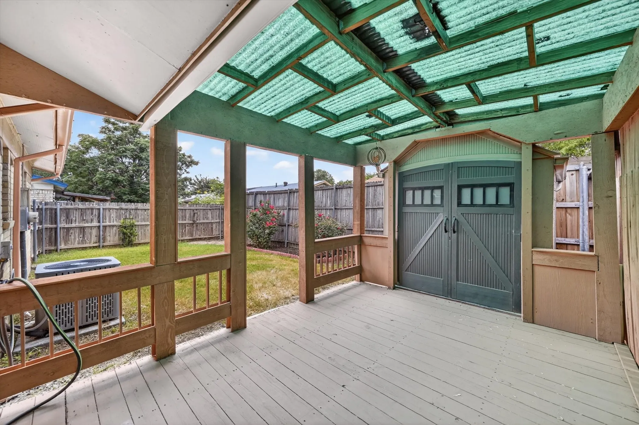 Covered wood-deck patio featuring a corrugated translucent roof, wood railings, and an integrated storage shed with double barn doors
