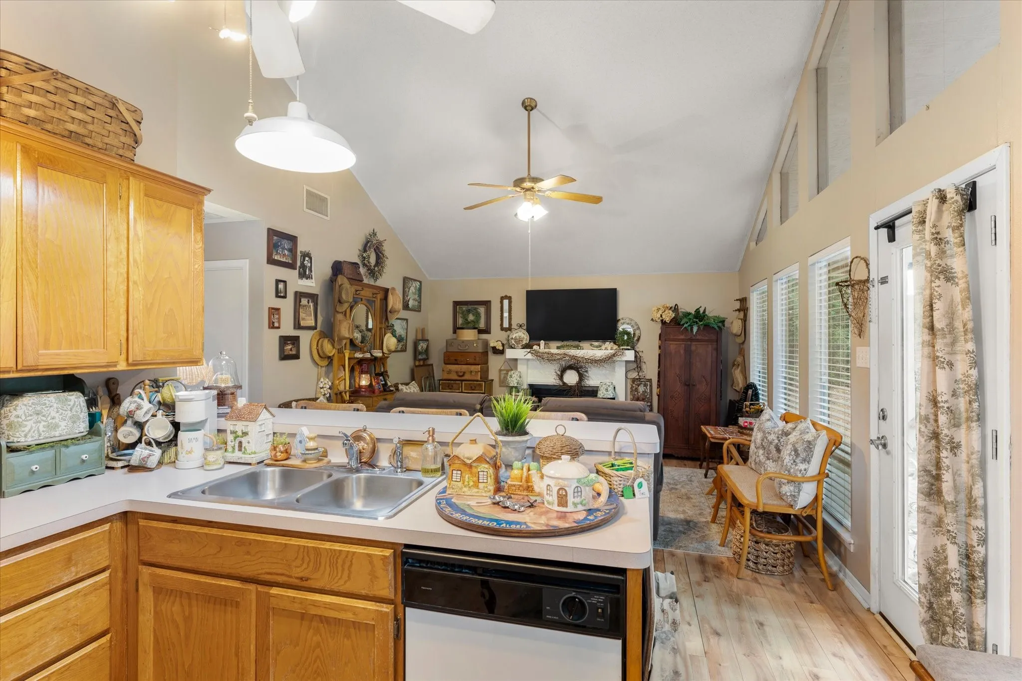 Open-concept kitchen and living area featuring vaulted ceilings, wood-finish flooring, ample natural light from multiple windows, a stainless steel double sink, and a built-in dishwasher