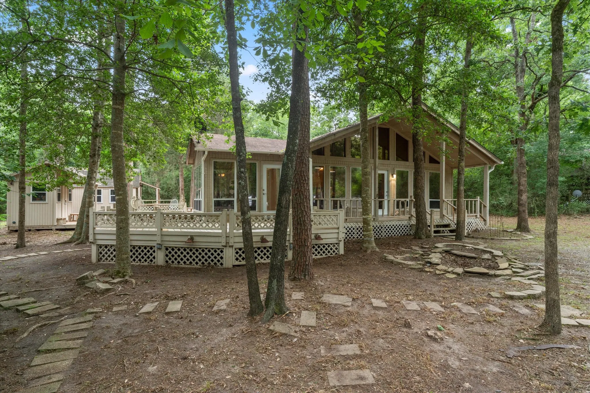Wooded property featuring a main residence with a prominent A-frame architectural style, a spacious exterior deck with lattice skirting, and an integrated stone fire pit area
