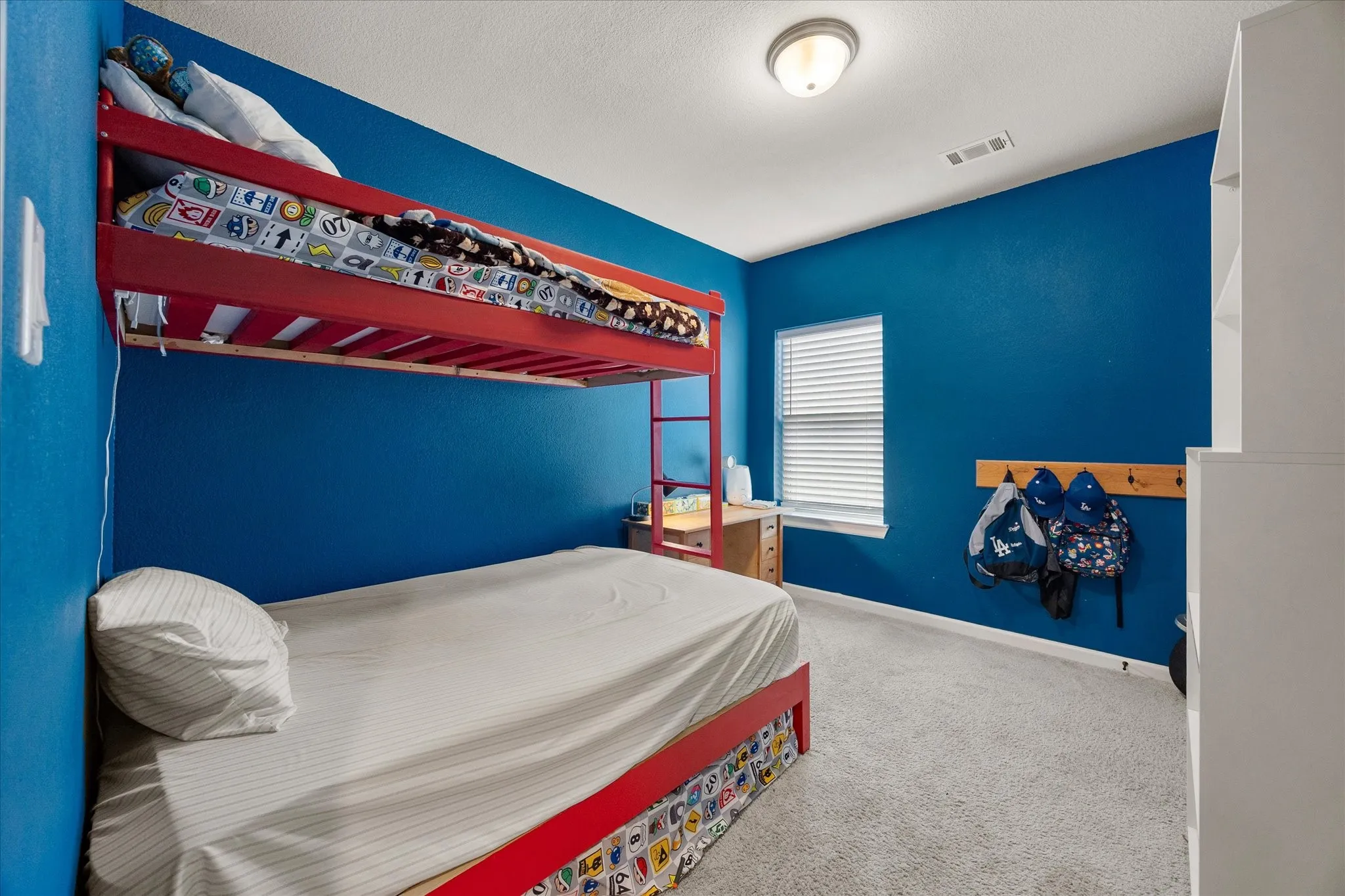 Bright bedroom featuring a single window with blinds, a built-in wooden coat rack, a white built-in shelving unit, and neutral carpet flooring
