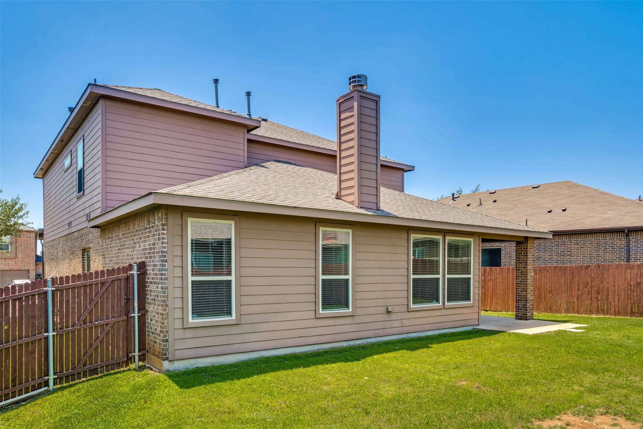 Expansive backyard featuring a brick and siding exterior, multiple windows, a prominent chimney, and a concrete patio