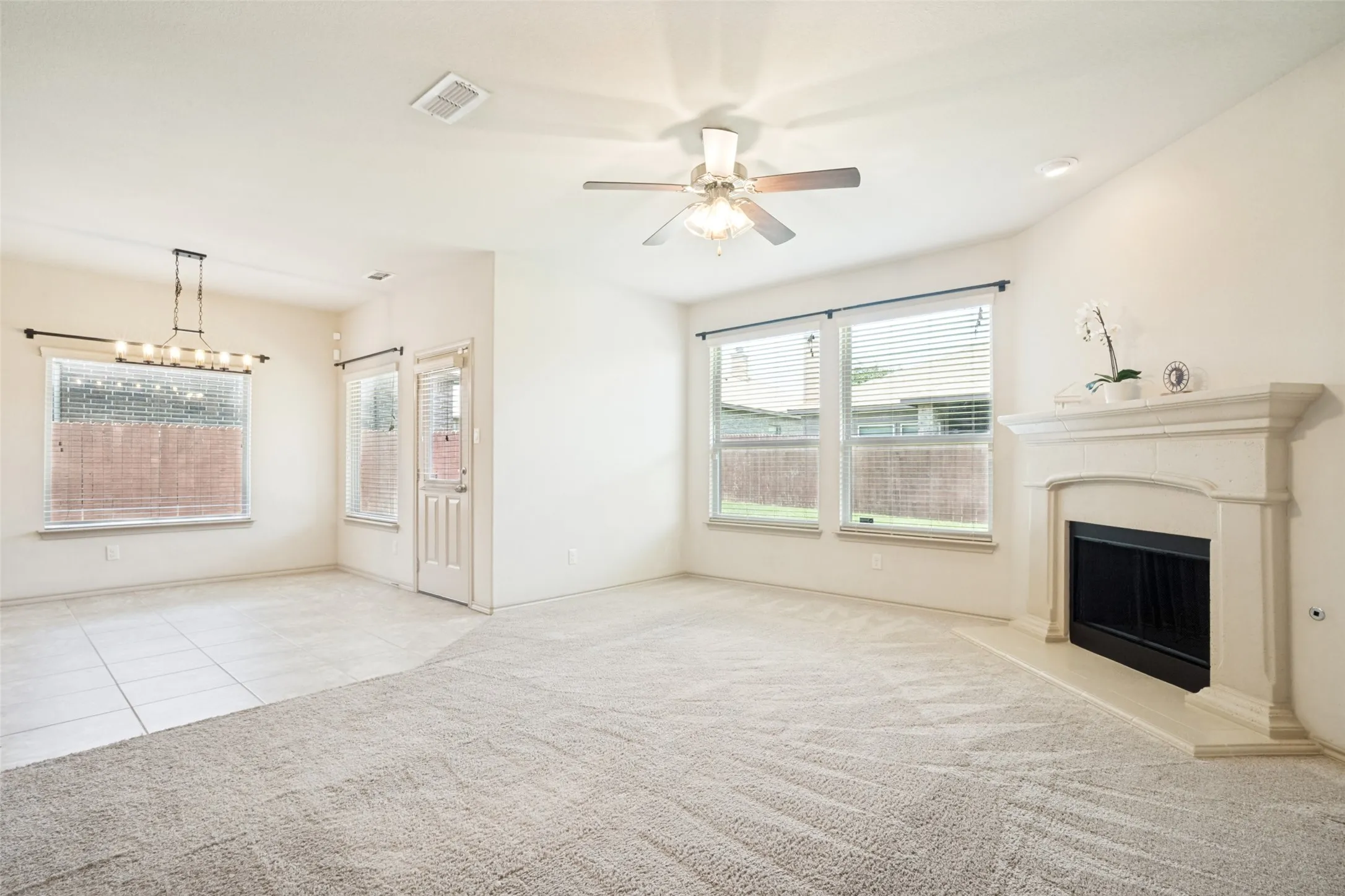 Spacious living area featuring a fireplace with a mantel, multiple windows with blinds, a ceiling fan, and carpet flooring transitioning to tile