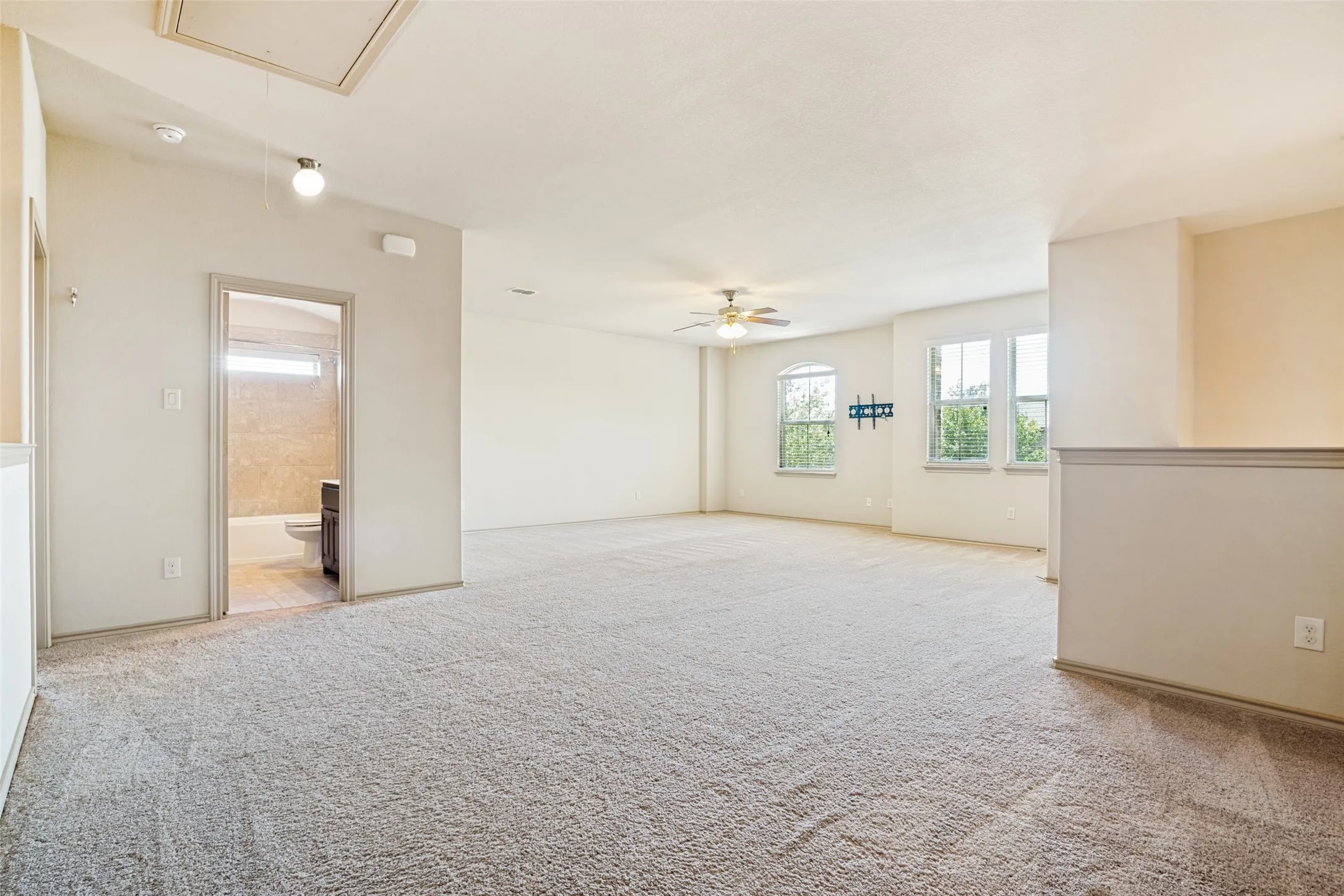 Spacious carpeted room featuring multiple windows, an overhead ceiling fan, and access to a bathroom with a white bathtub and dark wood-finish vanity