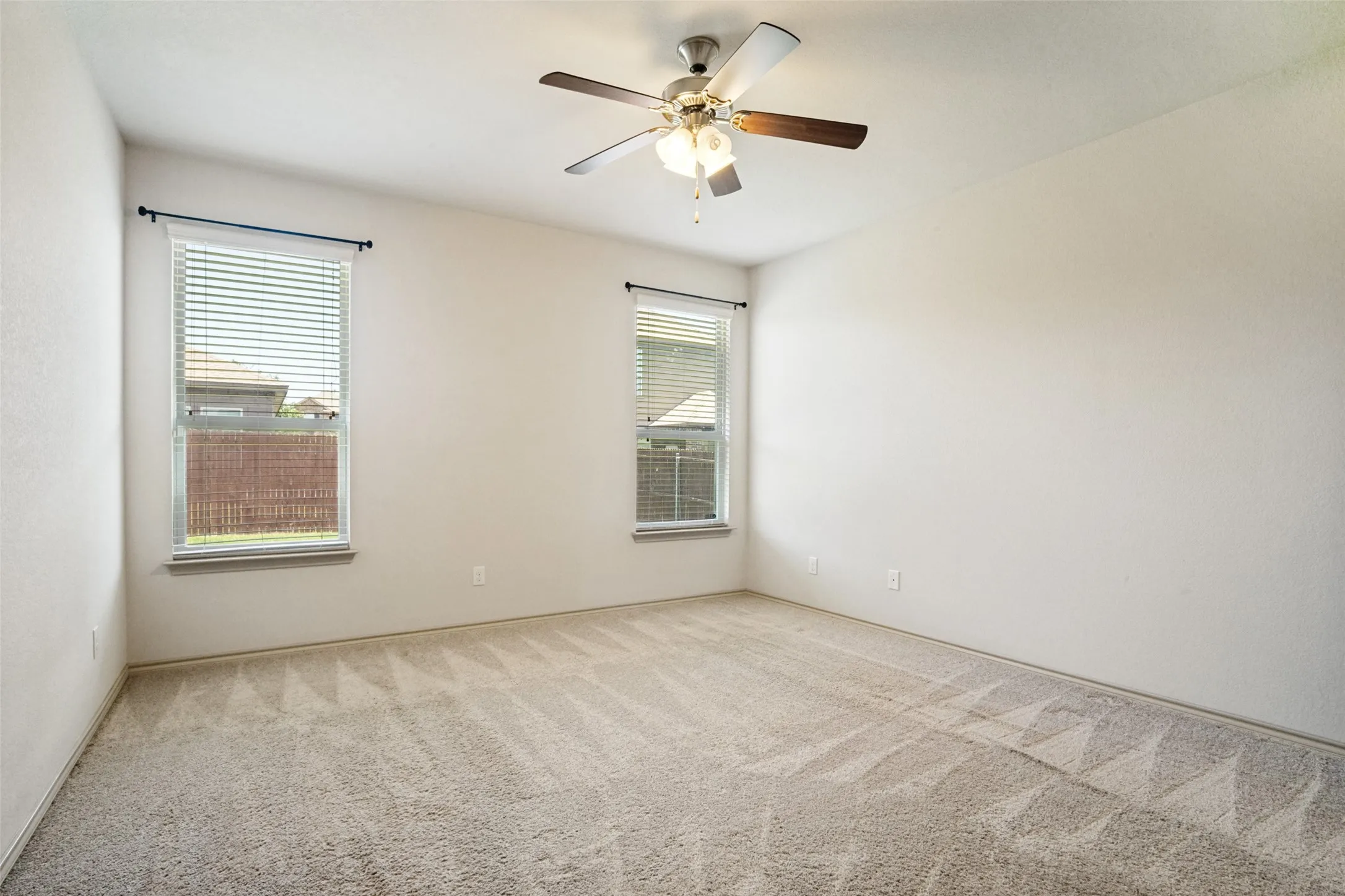 Spacious room featuring neutral carpet flooring, two windows with blinds, and a ceiling fan with integrated lighting