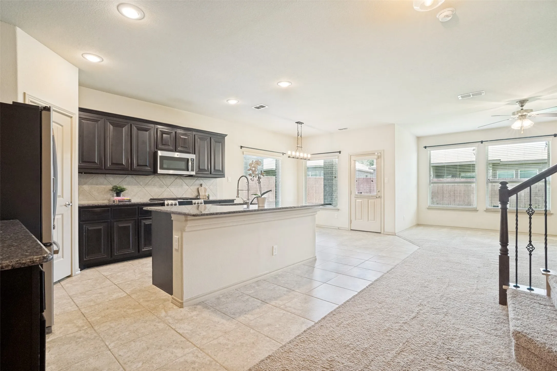 Open-concept living space with a kitchen island, dark cabinetry, tile flooring, and carpeted stairs with wrought iron balusters
