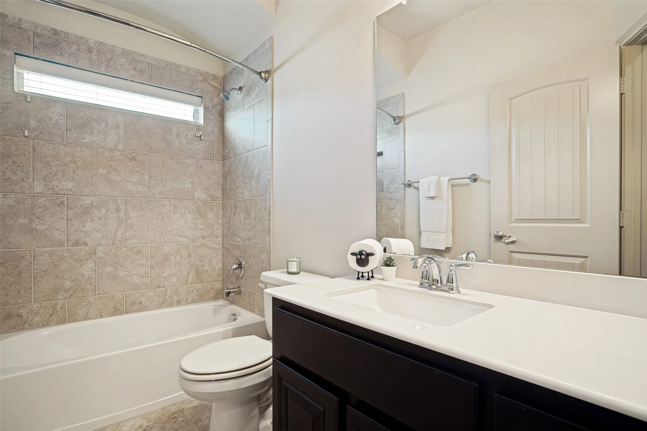 Bathroom featuring a dark wood-finish vanity with a white countertop and undermount sink, chrome fixtures, a full-width mirror, and a tub with light-toned tile surround and a transom window
