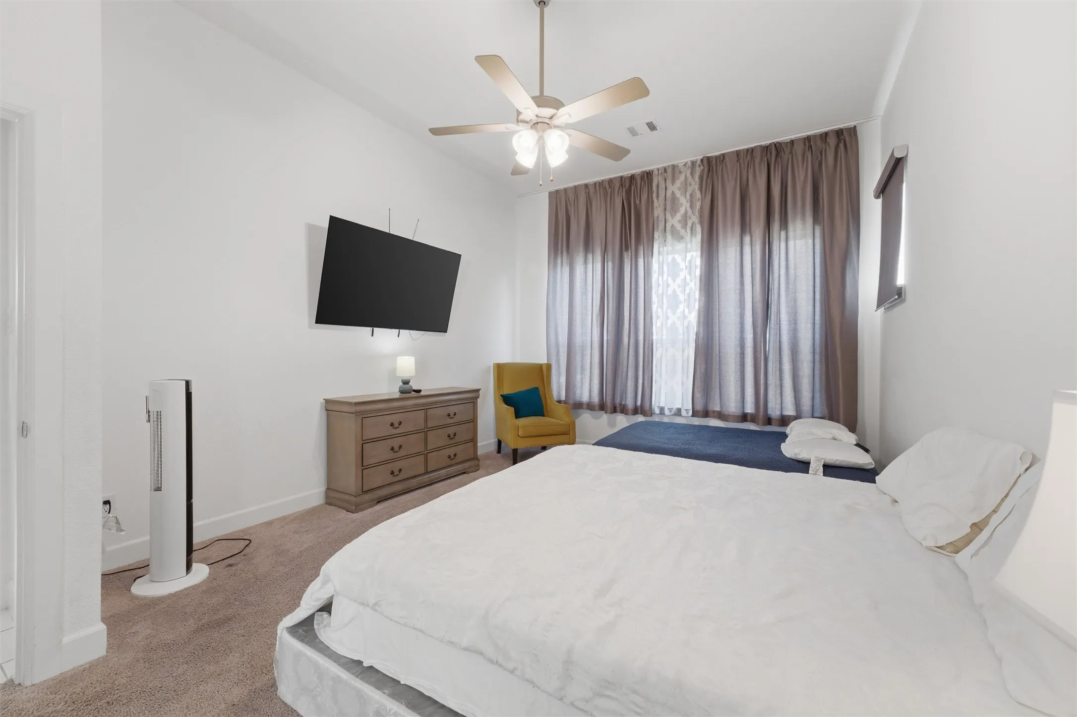 Carpeted room featuring a ceiling fan with integrated lighting, a wall-mounted television, and a window with both sheer and blackout curtains