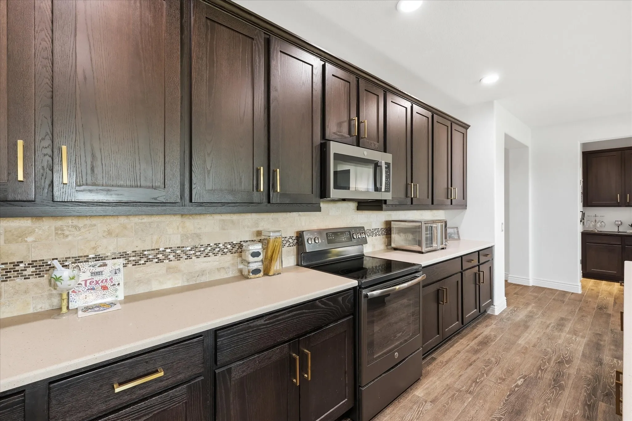 Kitchen featuring extensive dark wood-finish cabinetry with golden hardware, light-toned countertops, a tile backsplash with a decorative mosaic accent, and wood-finish flooring
