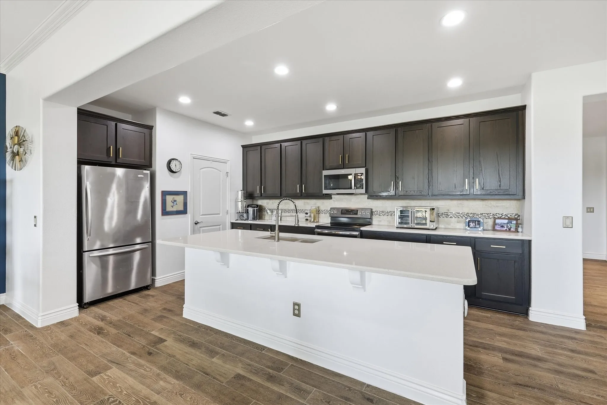 Modern kitchen featuring a large central island with a light-toned countertop, wood-finish flooring, dark-toned cabinetry, stainless steel appliances, and recessed lighting