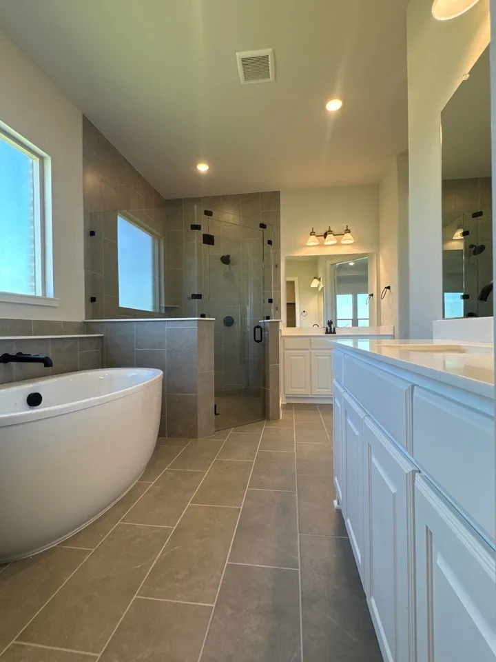 Freestanding soaking tub, glass-enclosed shower, dual vanity with white cabinetry, light-toned tile flooring, and recessed lighting