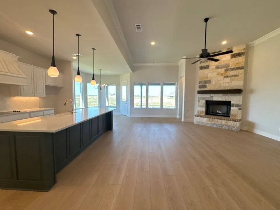 Spacious open-concept living area featuring wood-finish flooring, a stone-clad fireplace with a wood mantel, and a kitchen island with pendant lighting