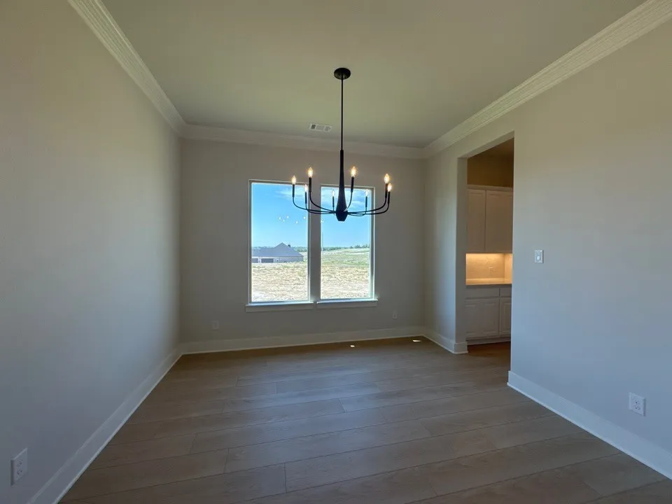 Expansive room featuring wood-look flooring, a contemporary chandelier, two large windows, crown molding, and an arched entry to a kitchen or pantry space with white cabinetry and under-cabinet lighting