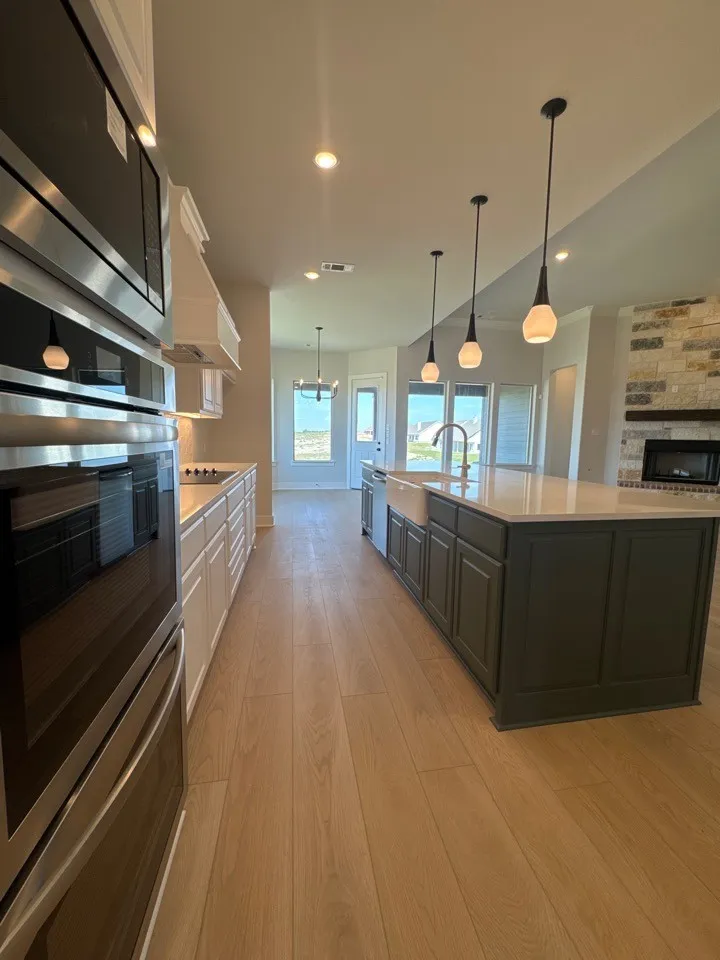 Built-in stainless steel appliances, wood-finish flooring, central island with pendant lighting, white farmhouse sink, and a stone-accented fireplace