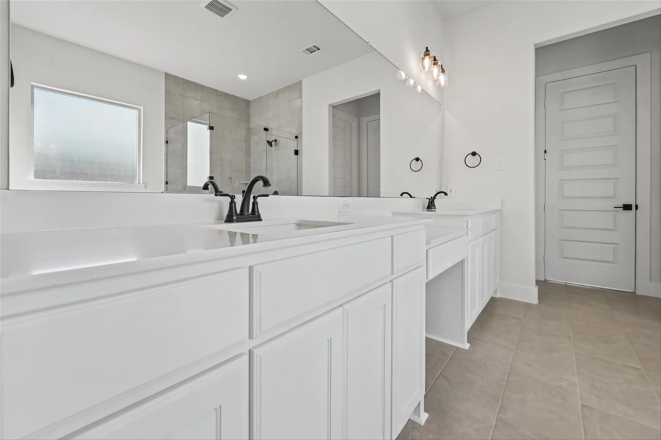 Dual white vanities with integrated sinks, matte black faucets, a large vanity mirror, and light-colored tile flooring