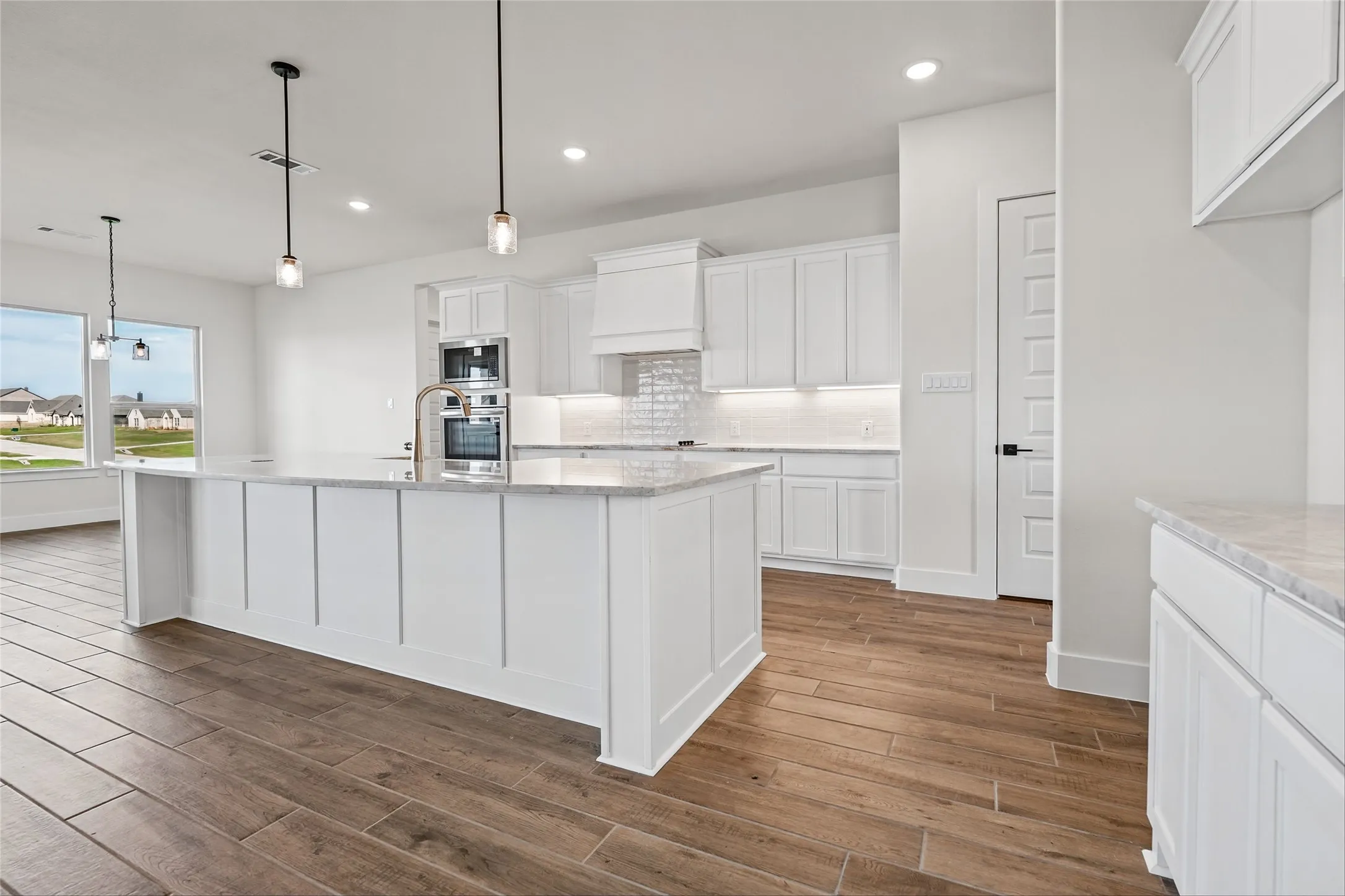 Kitchen featuring an expansive island with a sink, stainless steel built-in appliances, white shaker cabinetry, and wood-finish flooring