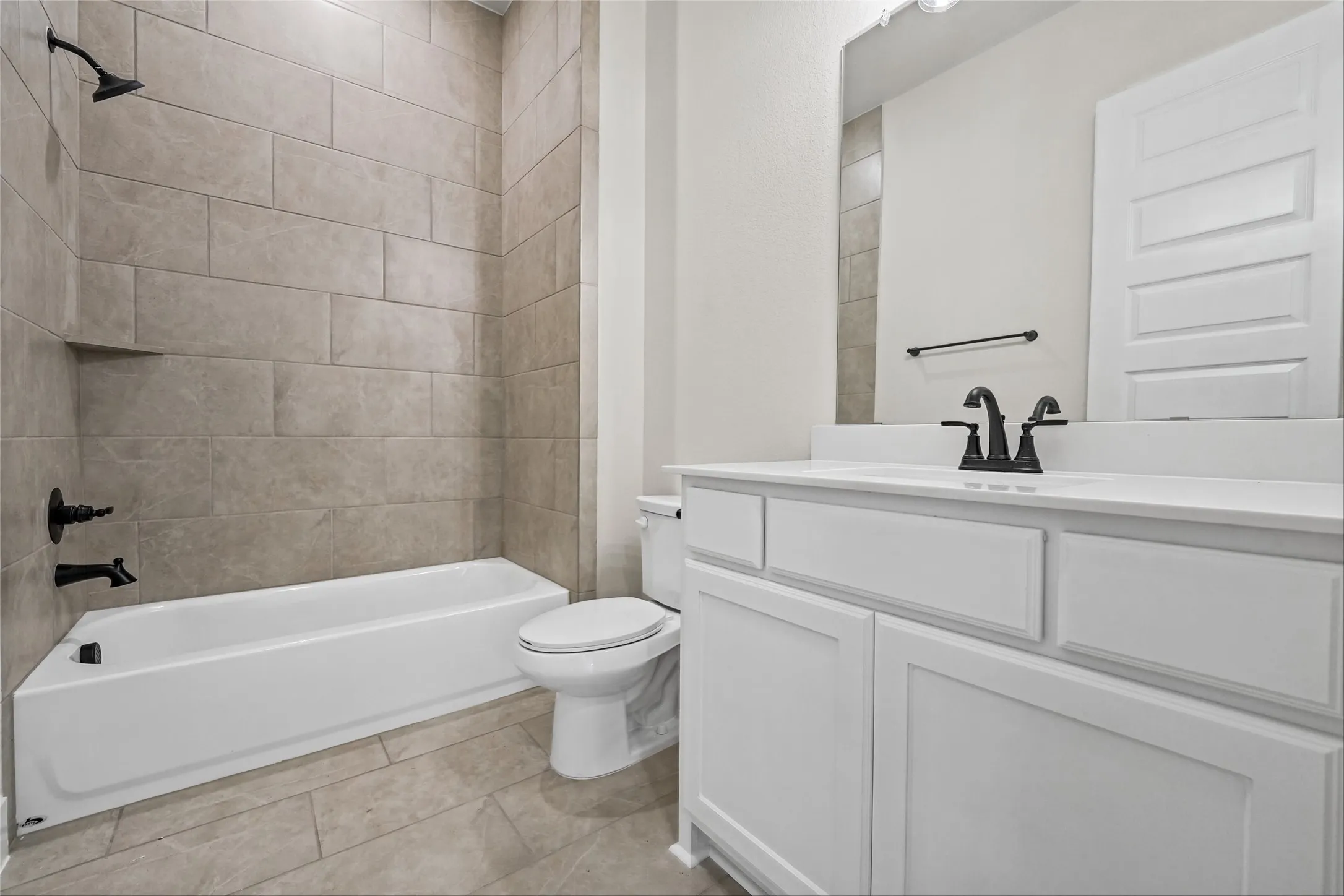 Bathtub with tiled surround and built-in niche, white vanity with integrated sink, black fixtures, and tiled flooring