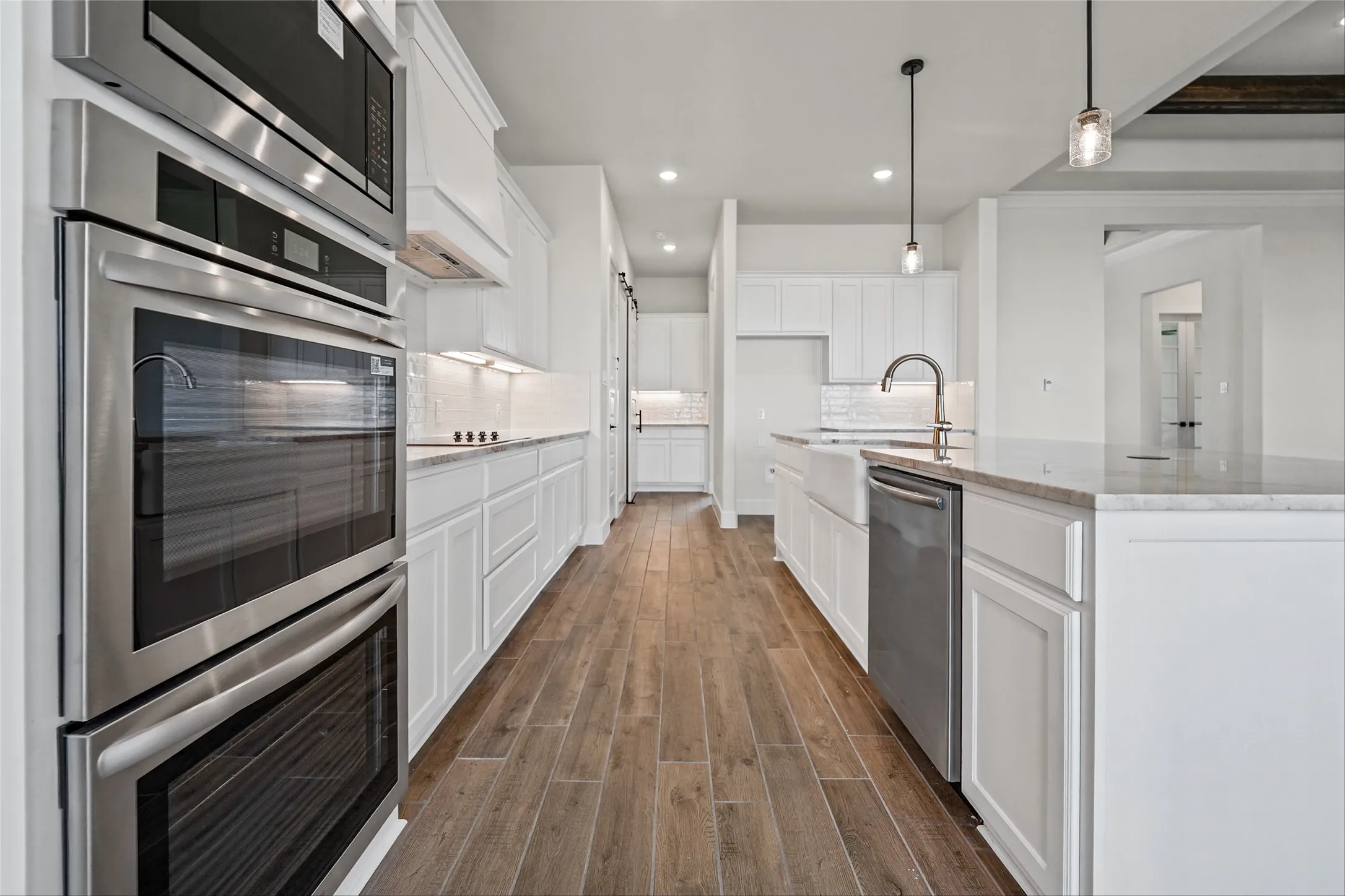 Kitchen featuring stainless steel double ovens, wood-finish flooring, white cabinetry, a farmhouse sink, and pendant lighting