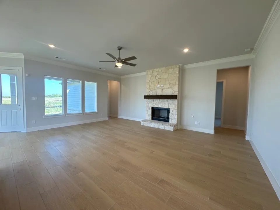 Spacious living area featuring wood-finish flooring, a stone-clad fireplace with a wood mantel, crown molding, a ceiling fan, and multiple windows