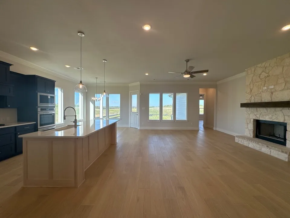Spacious open-concept interior featuring light wood-finish flooring, a stone fireplace with a wood-beam mantel, a kitchen island with pendant lighting, and built-in stainless steel appliances