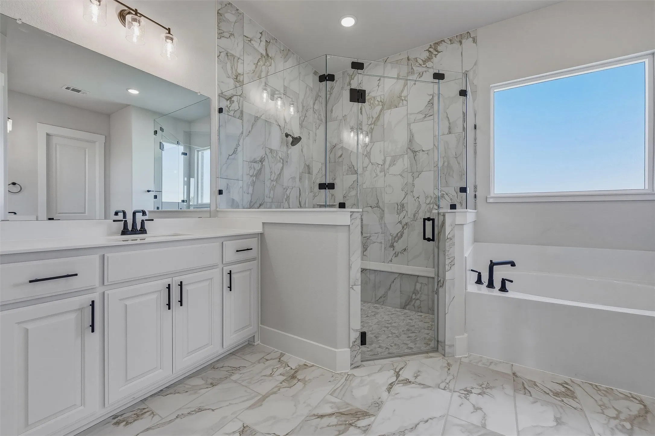 Dual vanity with white cabinetry and matte black hardware, spacious walk-in shower with glass enclosure and floor-to-ceiling tile, separate soaking tub with matte black fixtures, large window, and marble-look tile flooring