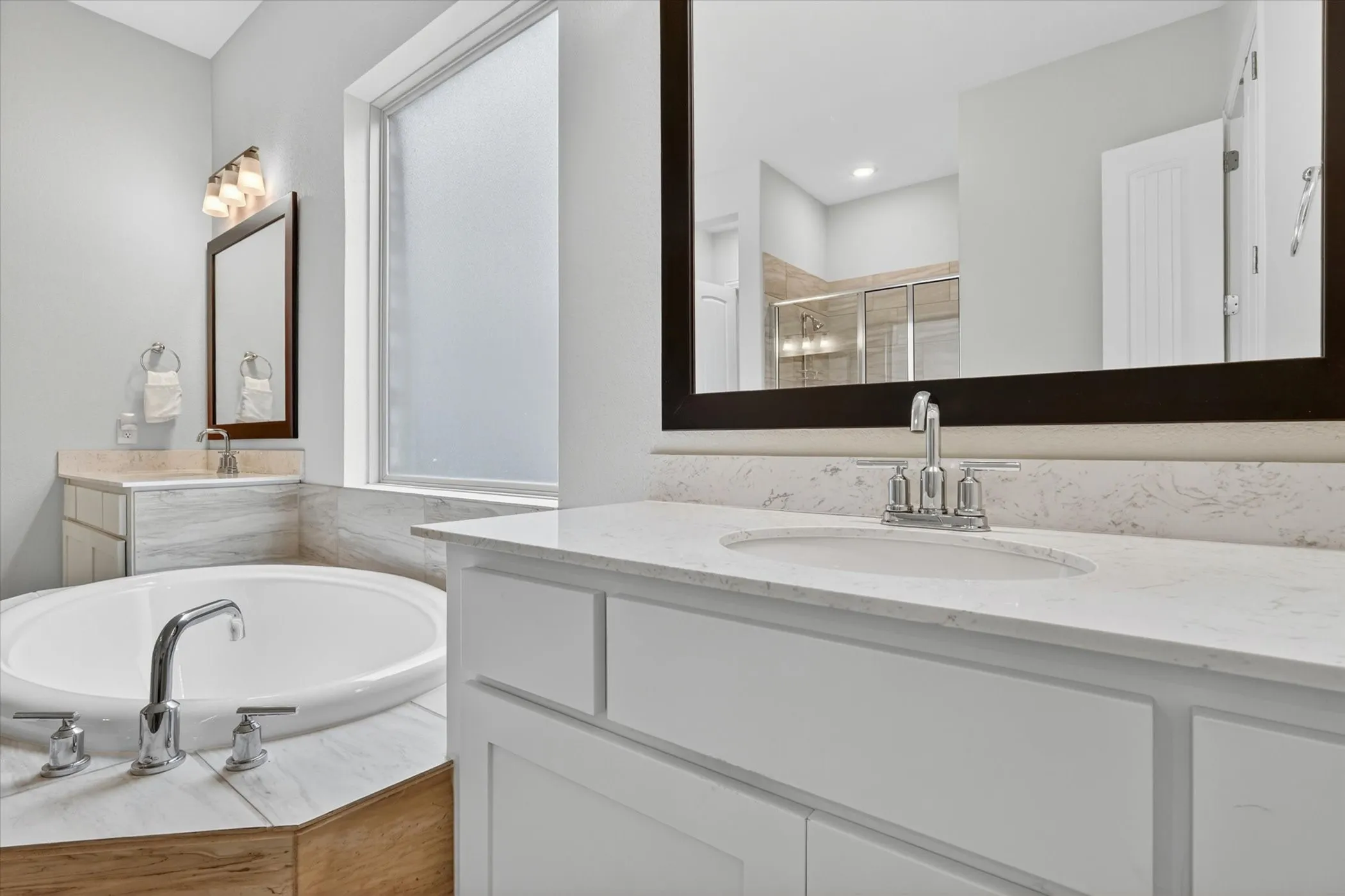 Spacious bathroom featuring a built-in soaking tub with chrome fixtures, dual vanities with white countertops, and a frosted privacy window