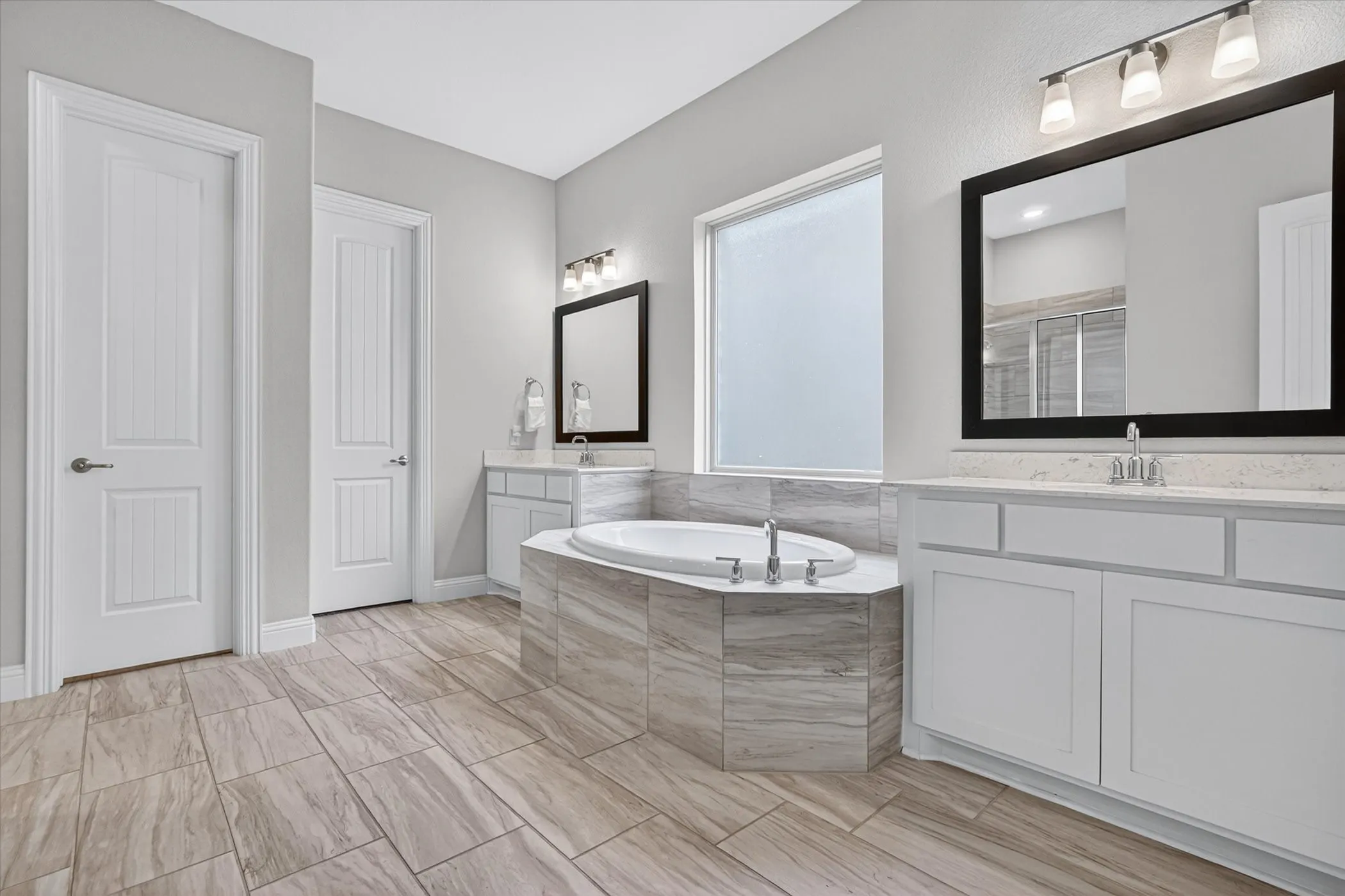 Spacious bathroom featuring a built-in soaking tub with surrounding tiled deck, large vanity with dual sinks, and ample natural light from a frosted window