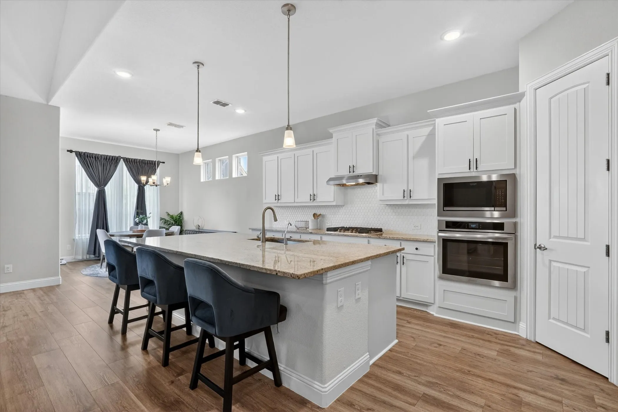 Open-concept kitchen featuring an island with stone countertops, white shaker cabinetry, stainless steel built-in appliances, a hexagonal tile backsplash, and wood-finish flooring