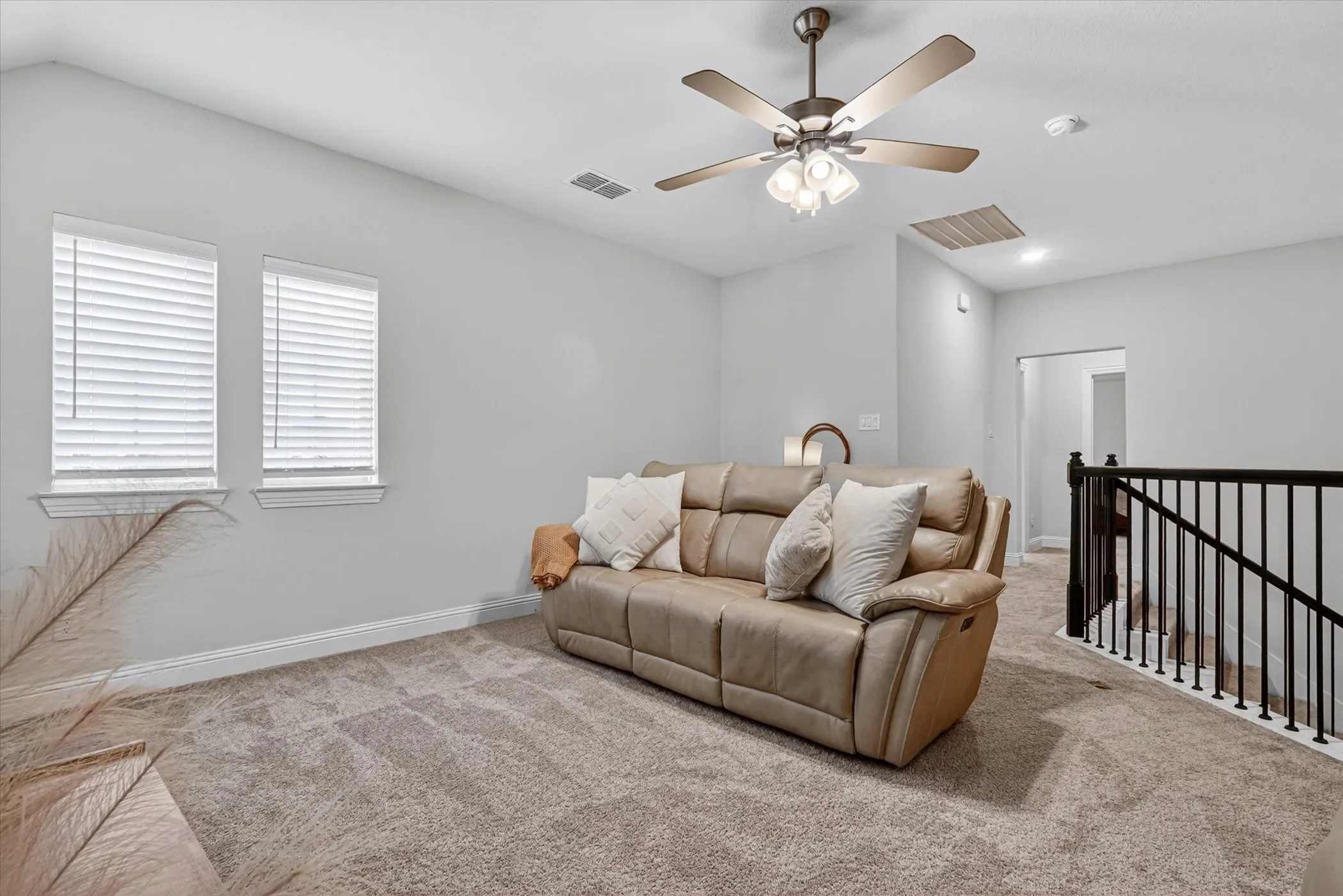 Carpeted bonus room featuring a ceiling fan with integrated lighting, two windows with blinds, and a black wrought iron staircase railing