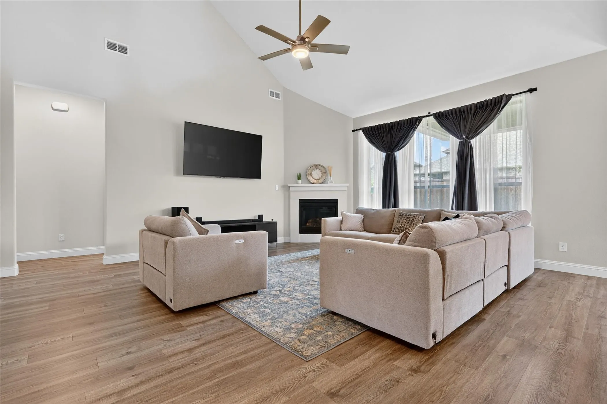 Spacious living area featuring vaulted ceilings, wood-finish flooring, a ceiling fan, a white mantel fireplace, and large windows