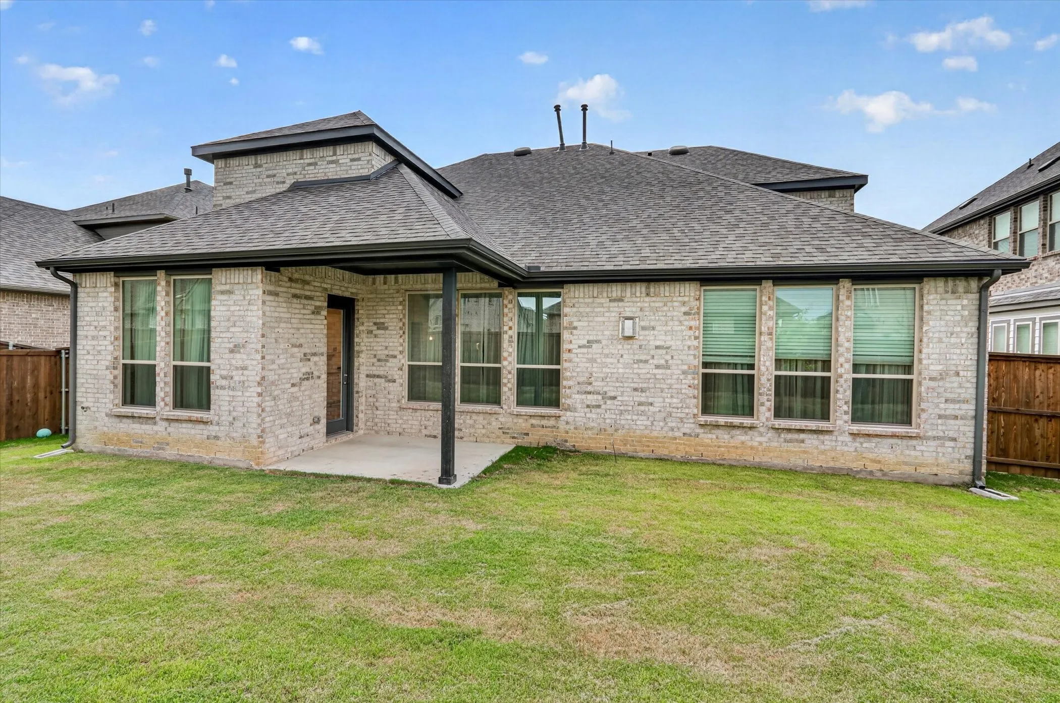 Expansive backyard featuring a manicured lawn, brick exterior, covered patio with concrete slab, and multiple large windows