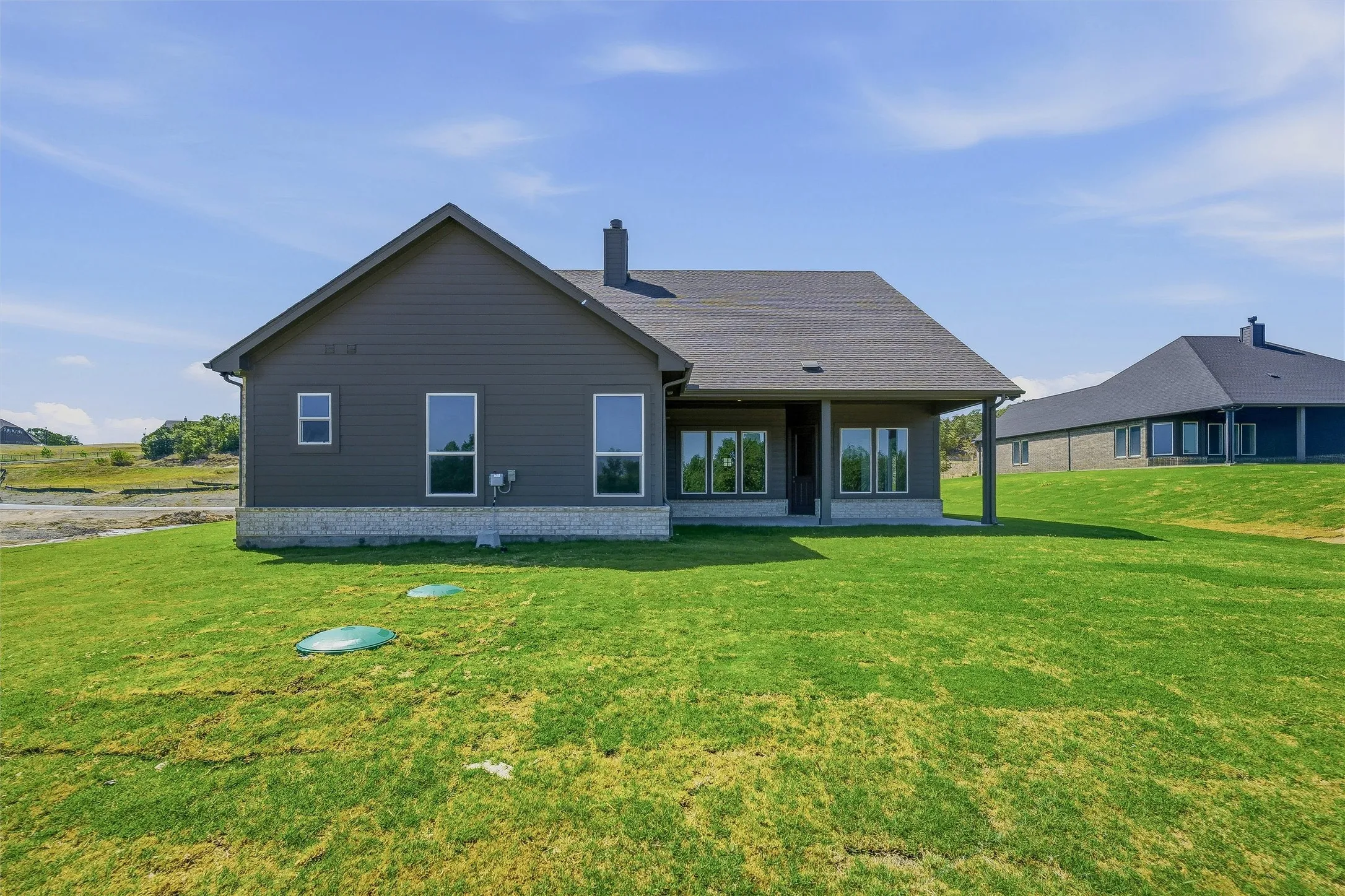 Contemporary residence featuring dark siding, a dark shingle roof, a stone-finish foundation, multiple large windows, and a covered patio
