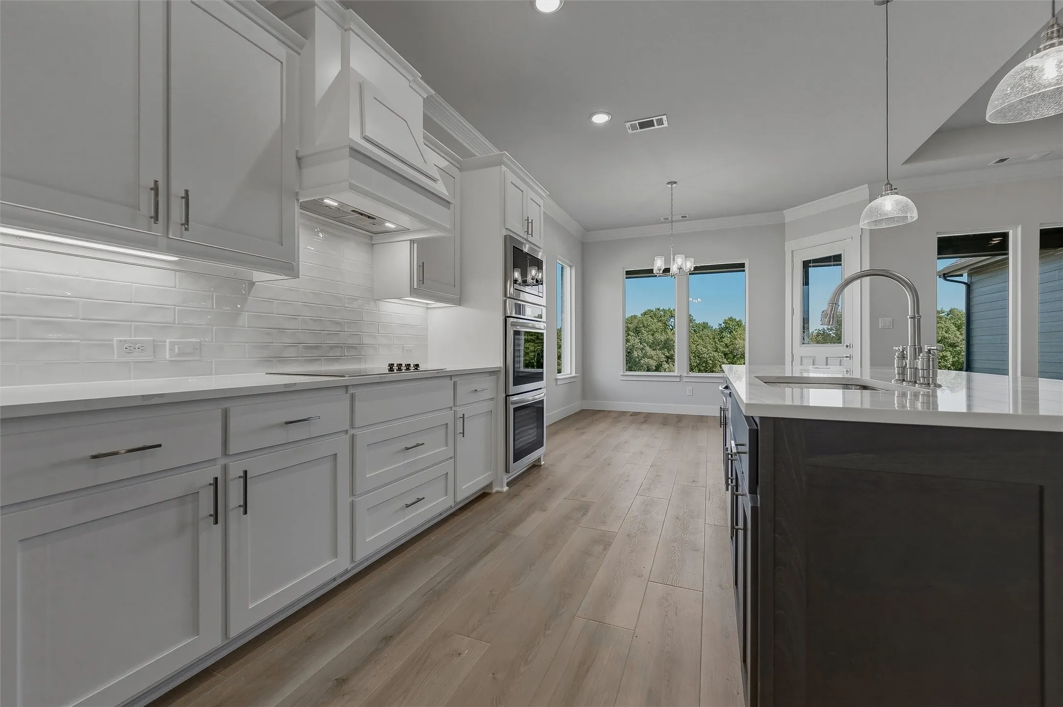 Kitchen featuring white cabinetry, light-toned wood-finish flooring, double wall ovens, a large center island with an undermount sink, and pendant lighting