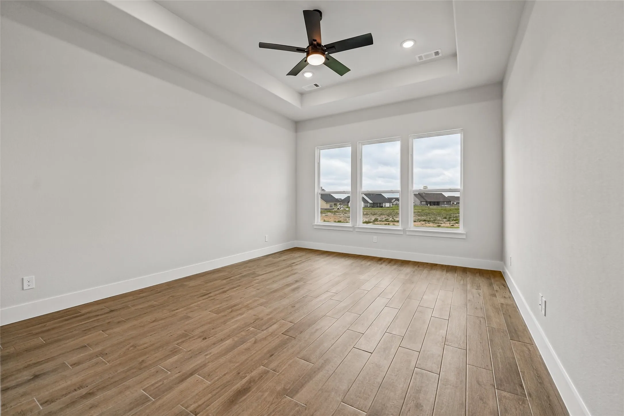 Tray ceiling with recessed lighting and a ceiling fan, three large windows, and wood-finish flooring