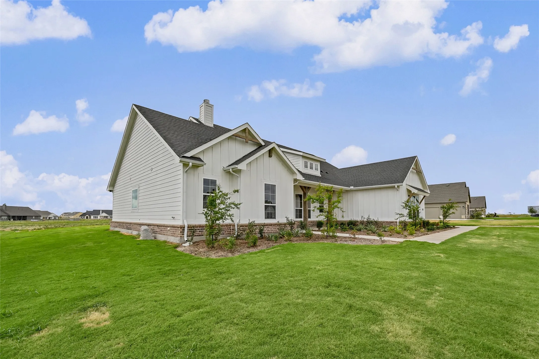 White siding exterior with dark shingle roofing, brick foundation, and established landscaping