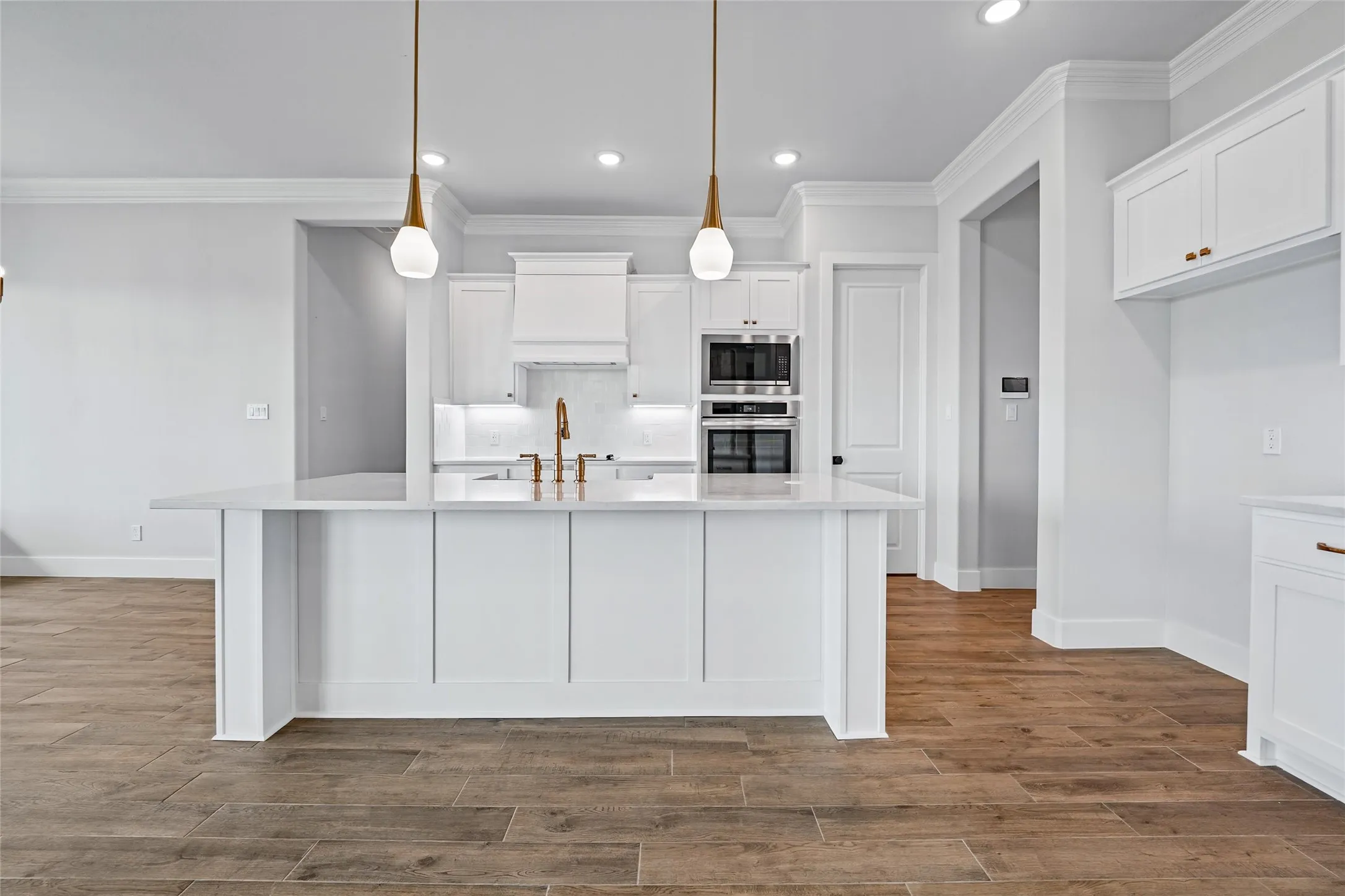 Kitchen island with a light-toned countertop, pendant lighting, stainless steel double ovens, a range hood, and wood-finish flooring