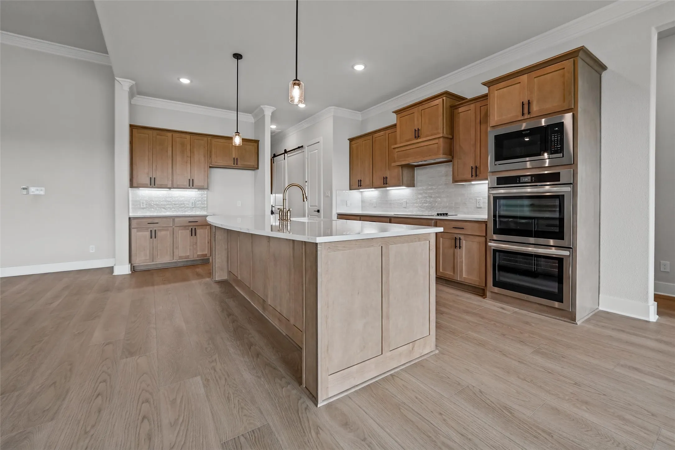 Spacious kitchen featuring an expansive center island with pendant lighting, wood-finish flooring, light-toned cabinetry, stainless steel built-in appliances, and a tiled backsplash