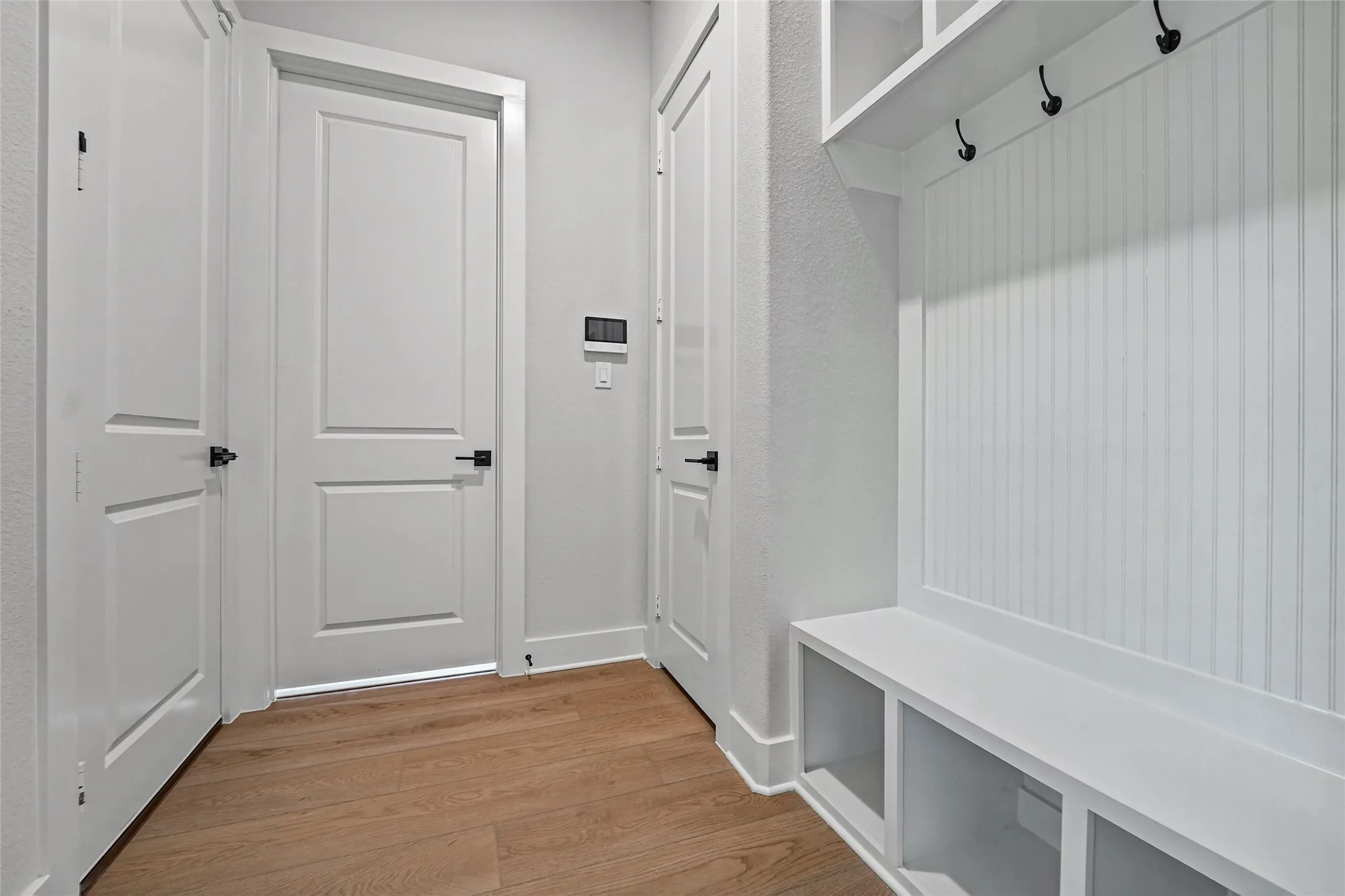 Wood-finish flooring, white panel doors with black hardware, and built-in white storage with shelving and coat hooks