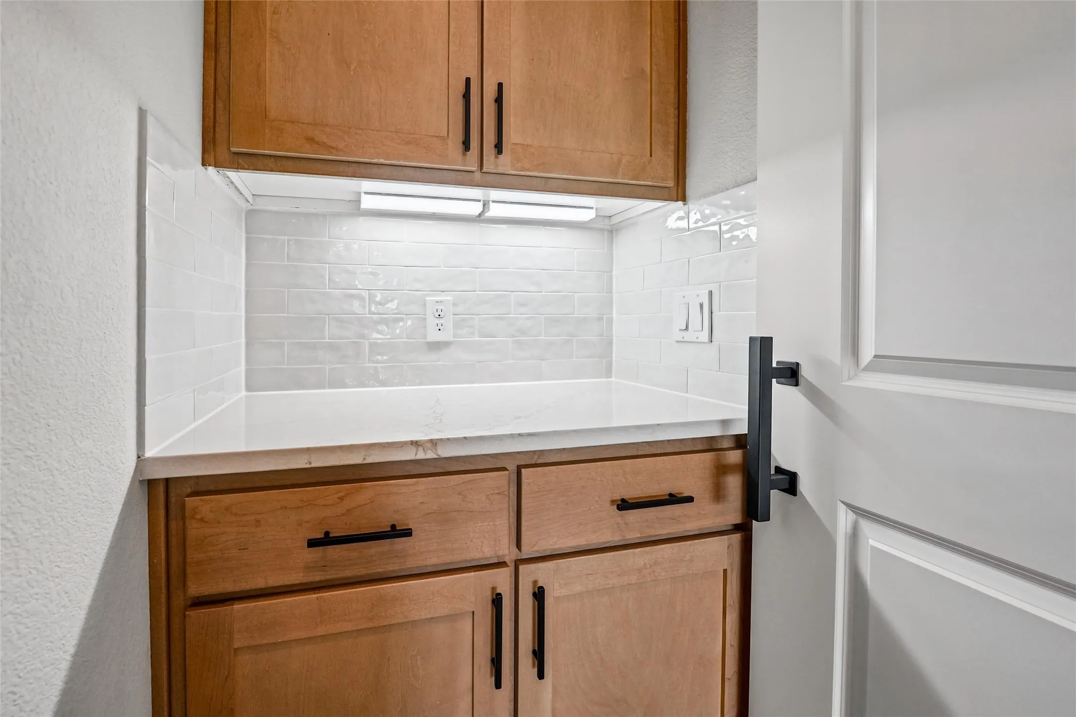 Built-in cabinetry with wood-finish doors and matte black hardware, white countertop, white subway tile backsplash, and under-cabinet lighting