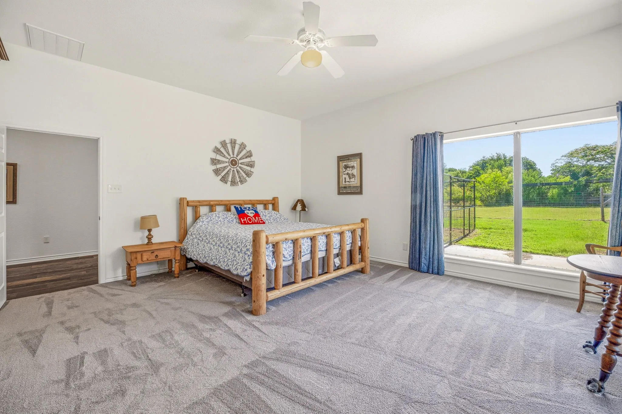 Spacious room featuring vaulted ceilings, a ceiling fan, plush gray carpeting, and a large picture window overlooking a fenced green space