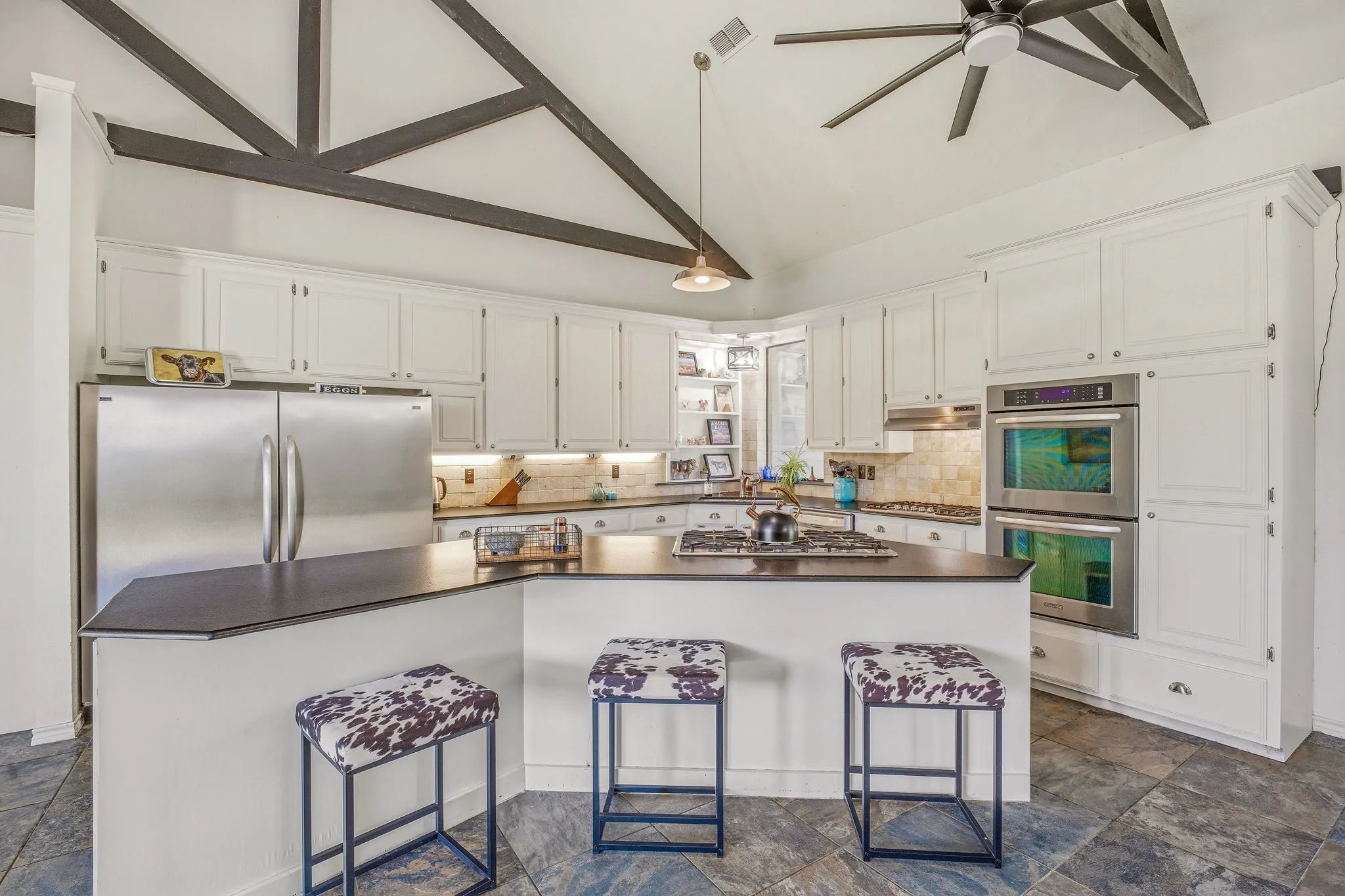 Kitchen featuring vaulted ceilings with exposed dark wood beams, white cabinetry, a large center island with a gas cooktop, stainless steel double ovens, and a tile backsplash